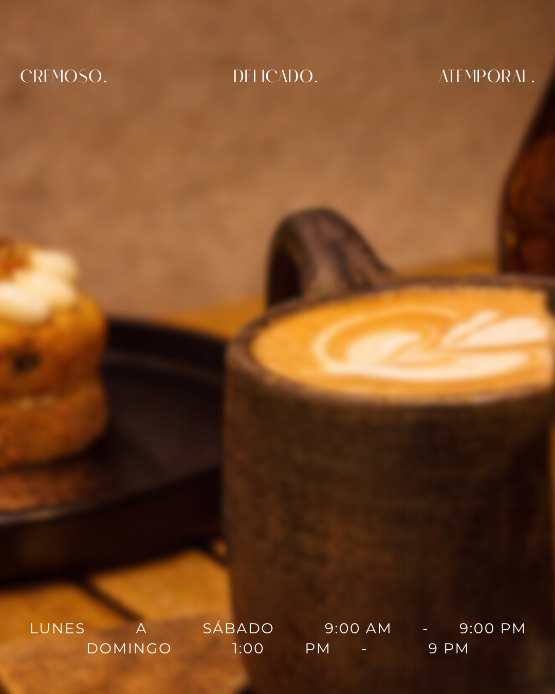 Una taza de café con leche decorada en la superficie y unos muffins en un plato negro, todo sobre una mesa de madera.