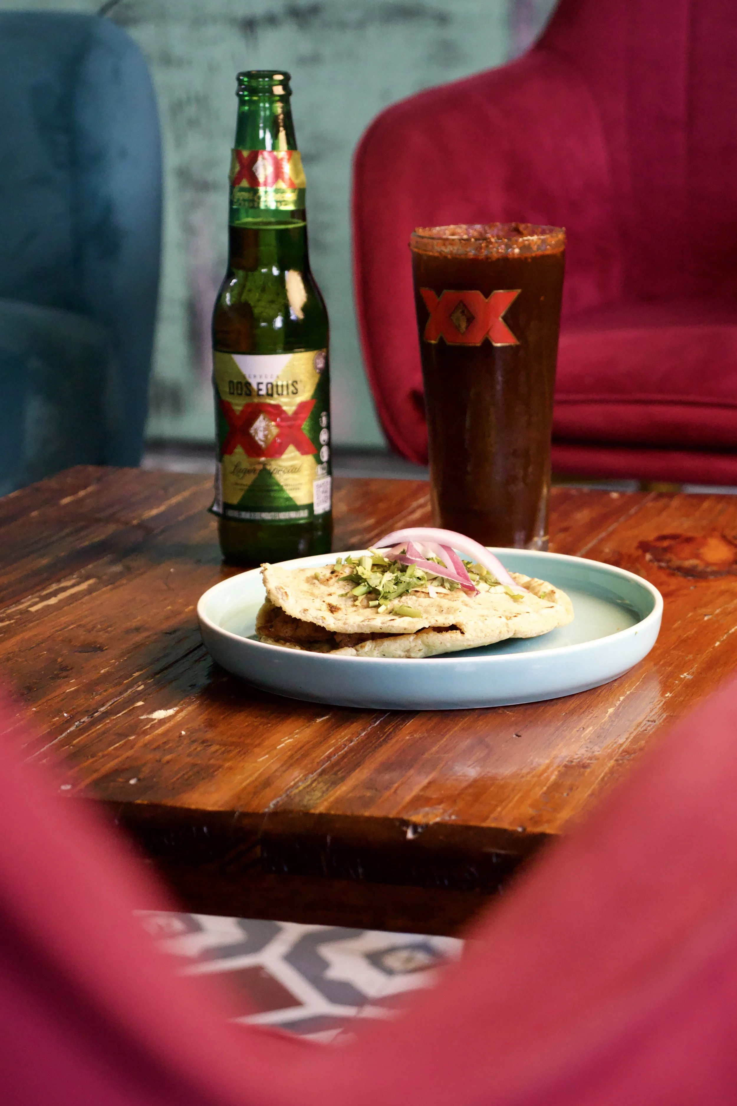 Una botella de cerveza Dos Equis, un vaso con cerveza oscura, y un plato con tacos y cebolla en un mesa de madera.