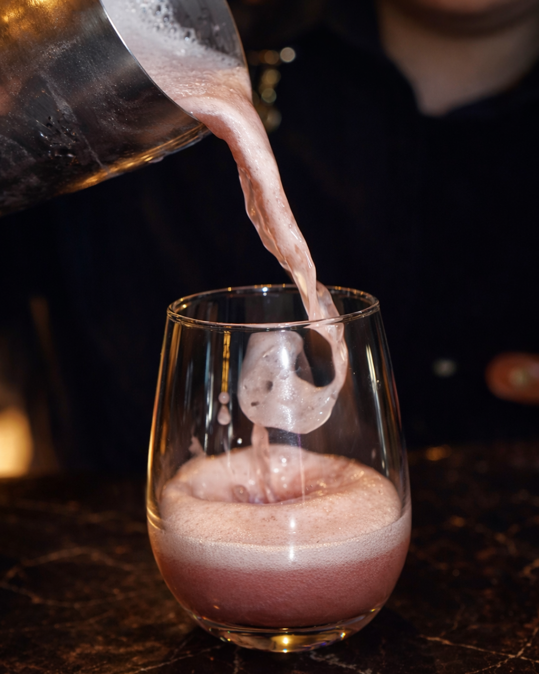 Vaso de cristal recibe una bebida rosada espumosa de una jarra de metal