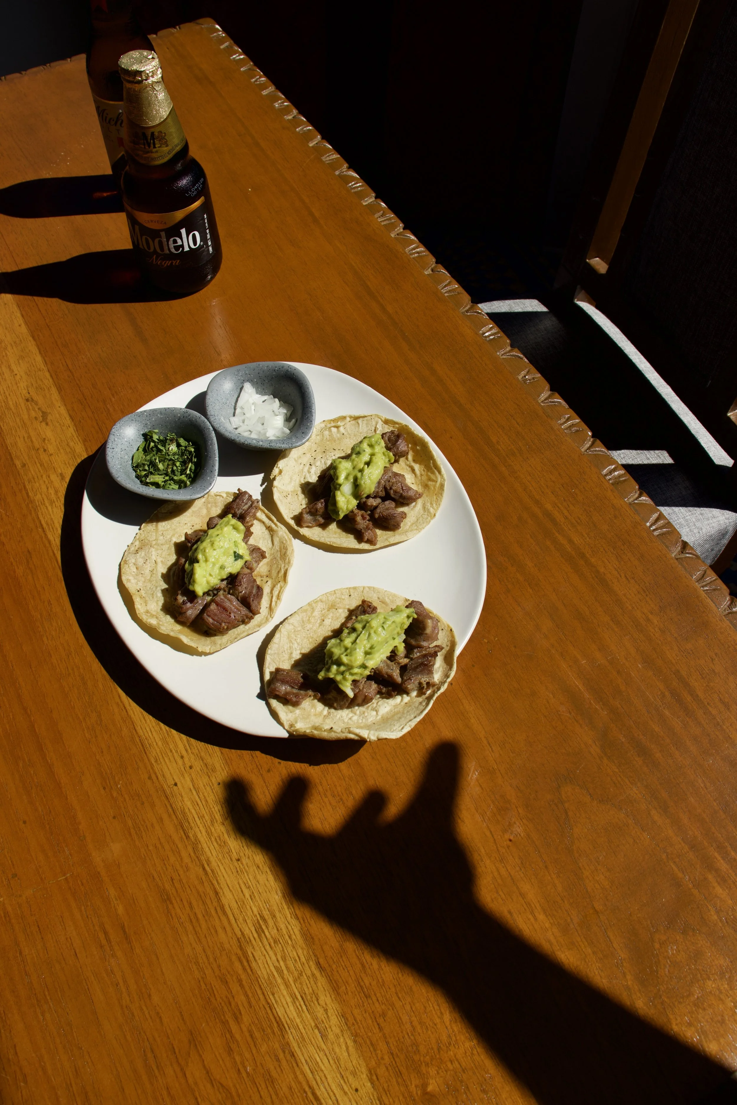 Tres tacos con carne, guacamole, y cebolla en un plato blanco sobre una mesa de madera.