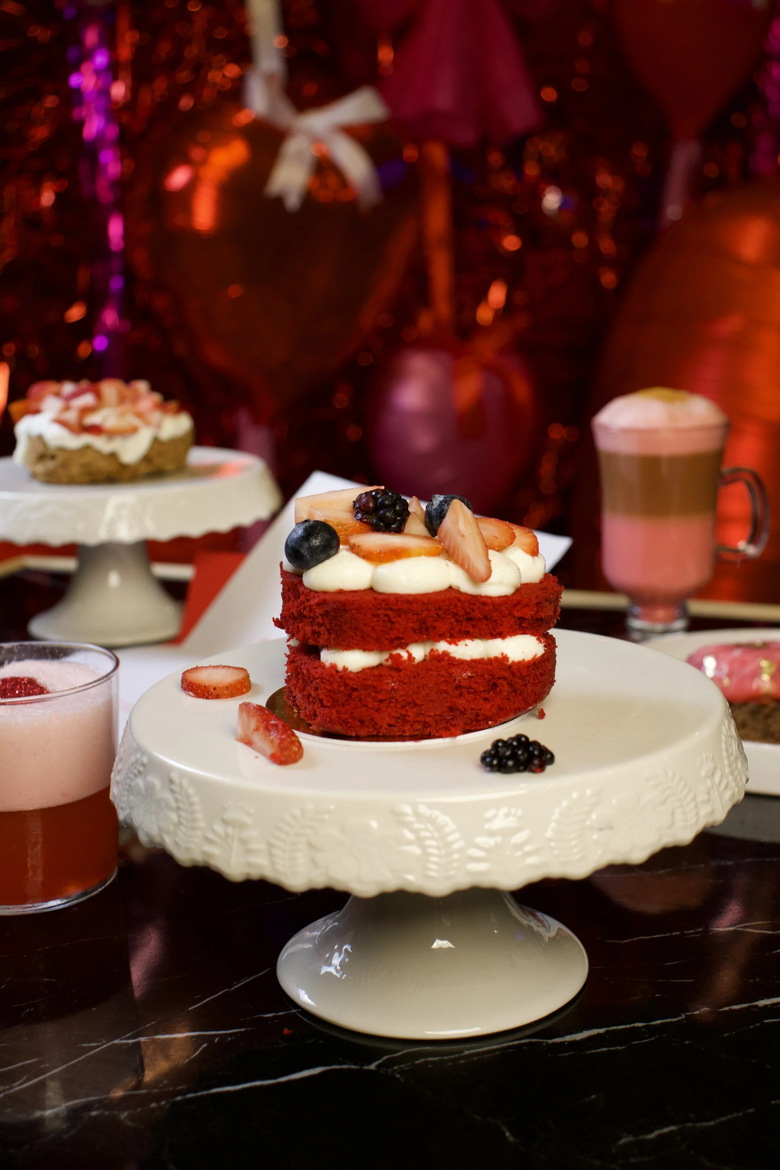 Pastel de red velvet con frosting blanco y frutas variadas en un plato decorativo blanco, rodeado de otros postres y bebidas en un ambiente festivo con decoraciones rojas y rosas.