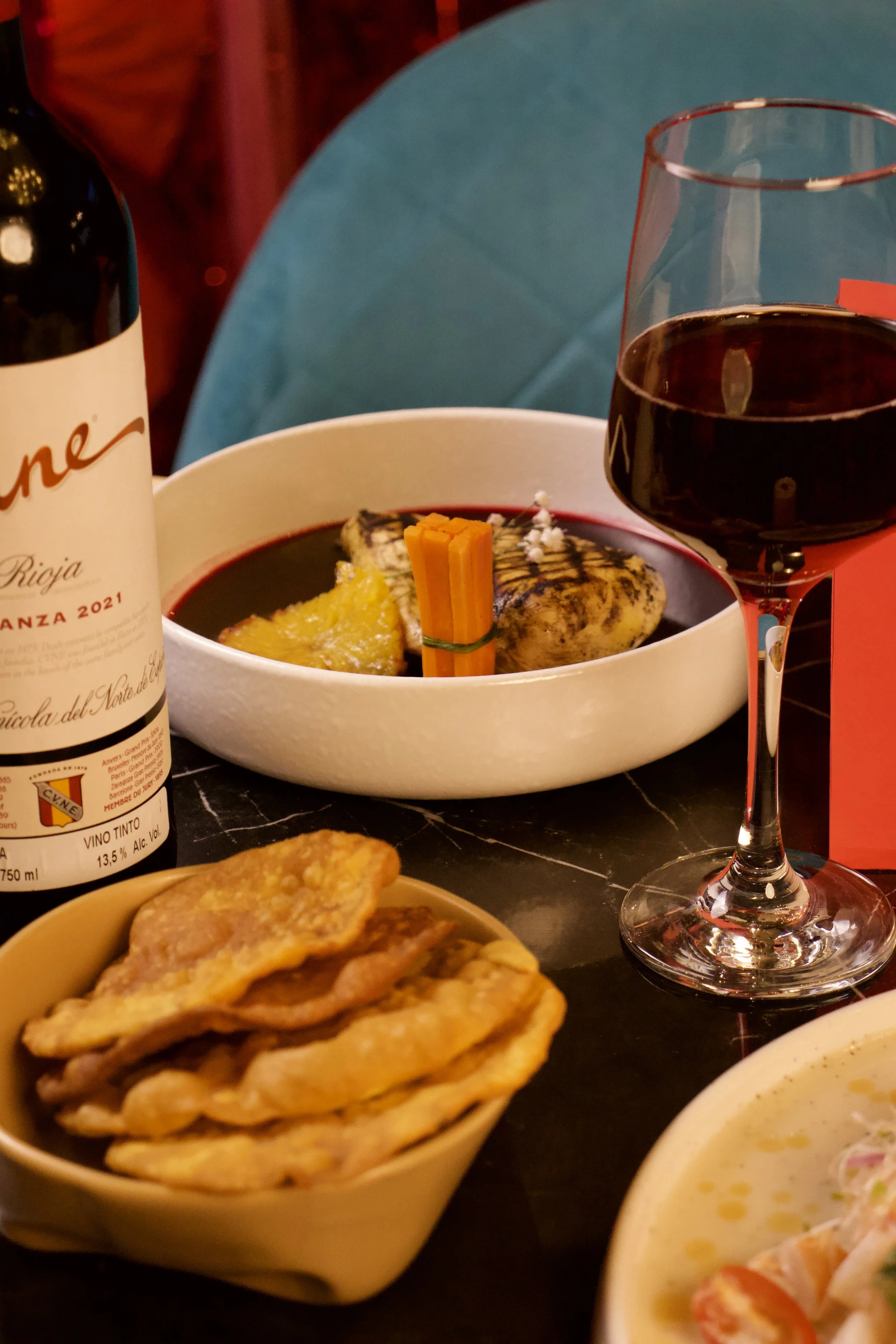 Plato de comida con vino tinto y una copa de vino, junto a un plato con pollo y vegetales, y una pequeña bowl con totopos.