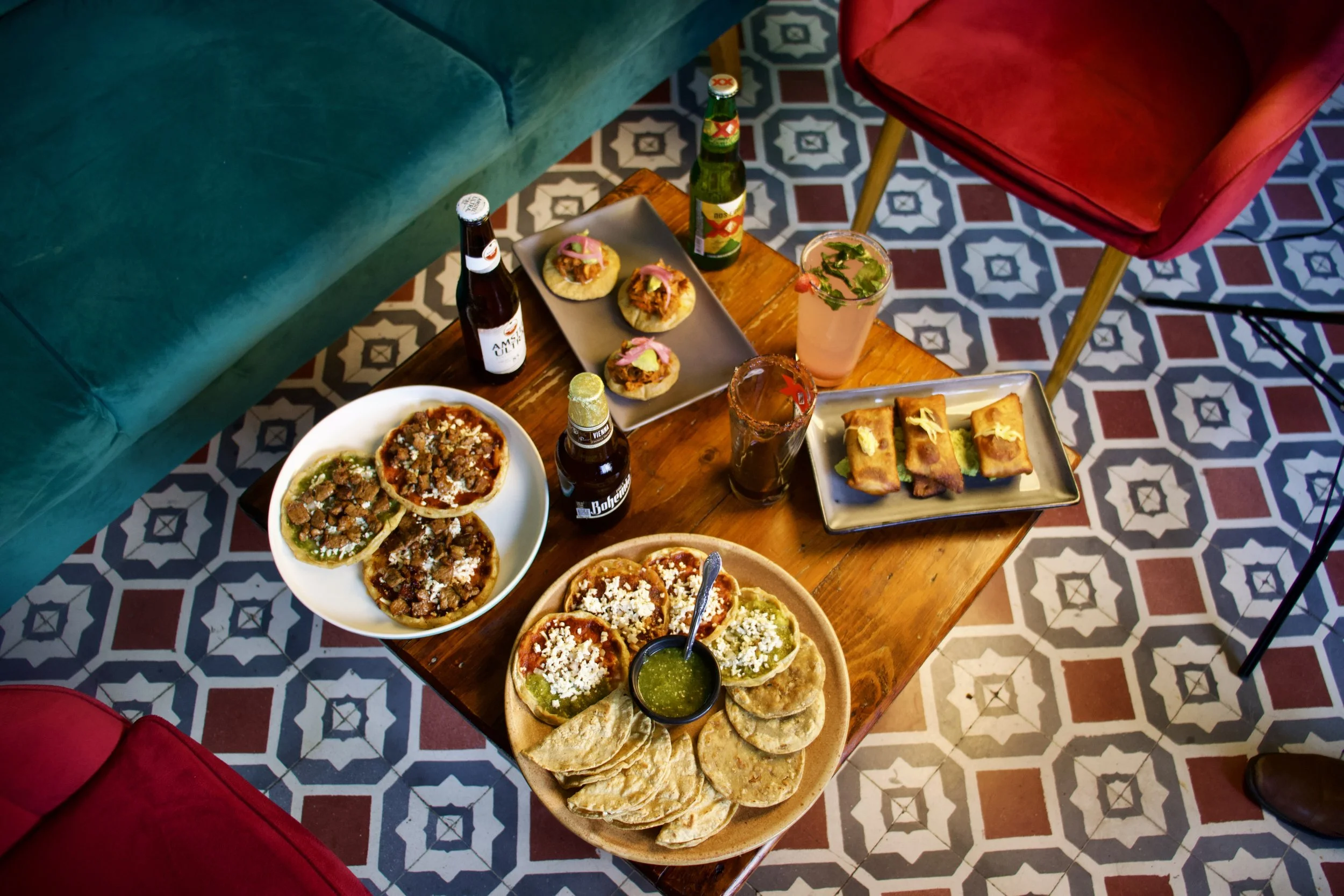 Mesa con comida mexicana, incluyendo tacos con salsas, totopos con guacamole, mini tacos, y bebidas. Rodeada por sillones de colores y un piso con mosaico.