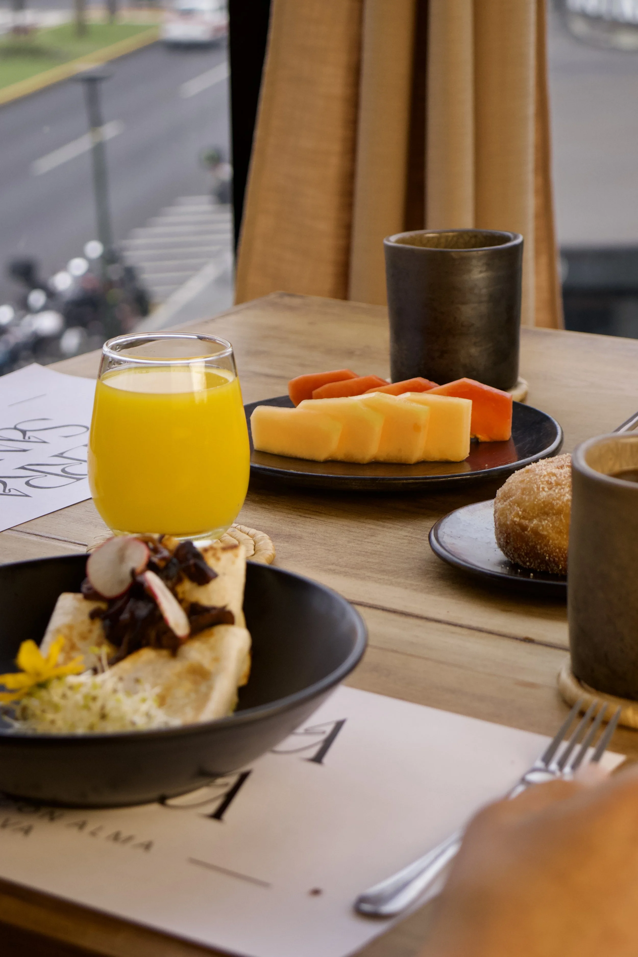 Mesa con desayuno que incluye jugo de naranja, fruta melón y papaya, un bol de cerdo con ensalada y torta, y dos tazas de incienso. Al fondo, una ventana con cortinas amarillas y vista a la calle.