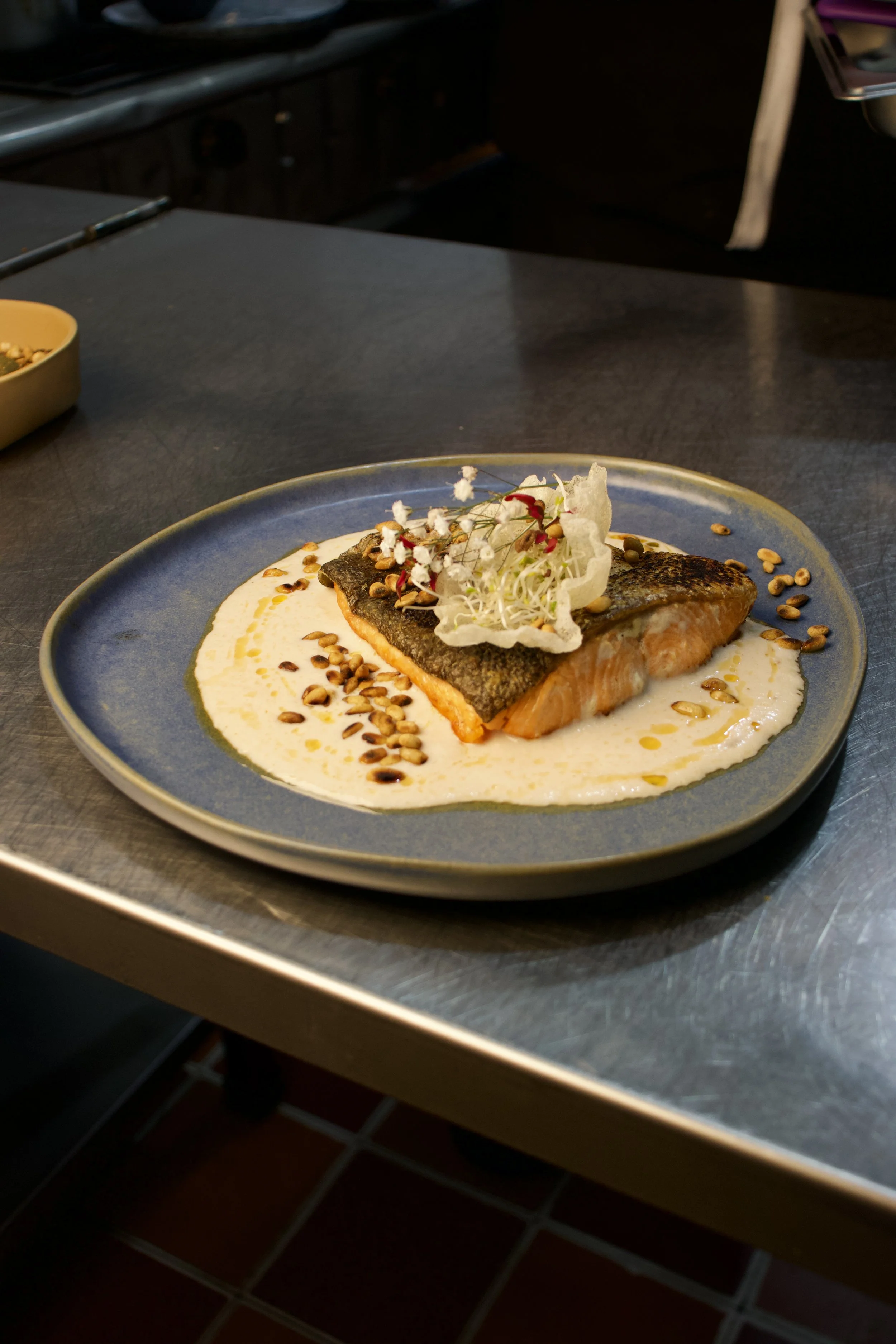 Filete de salmón con piel, servido en salsa blanca, adornado con brotes y semillas, en un plato azul. Fondo difuso de cocina.