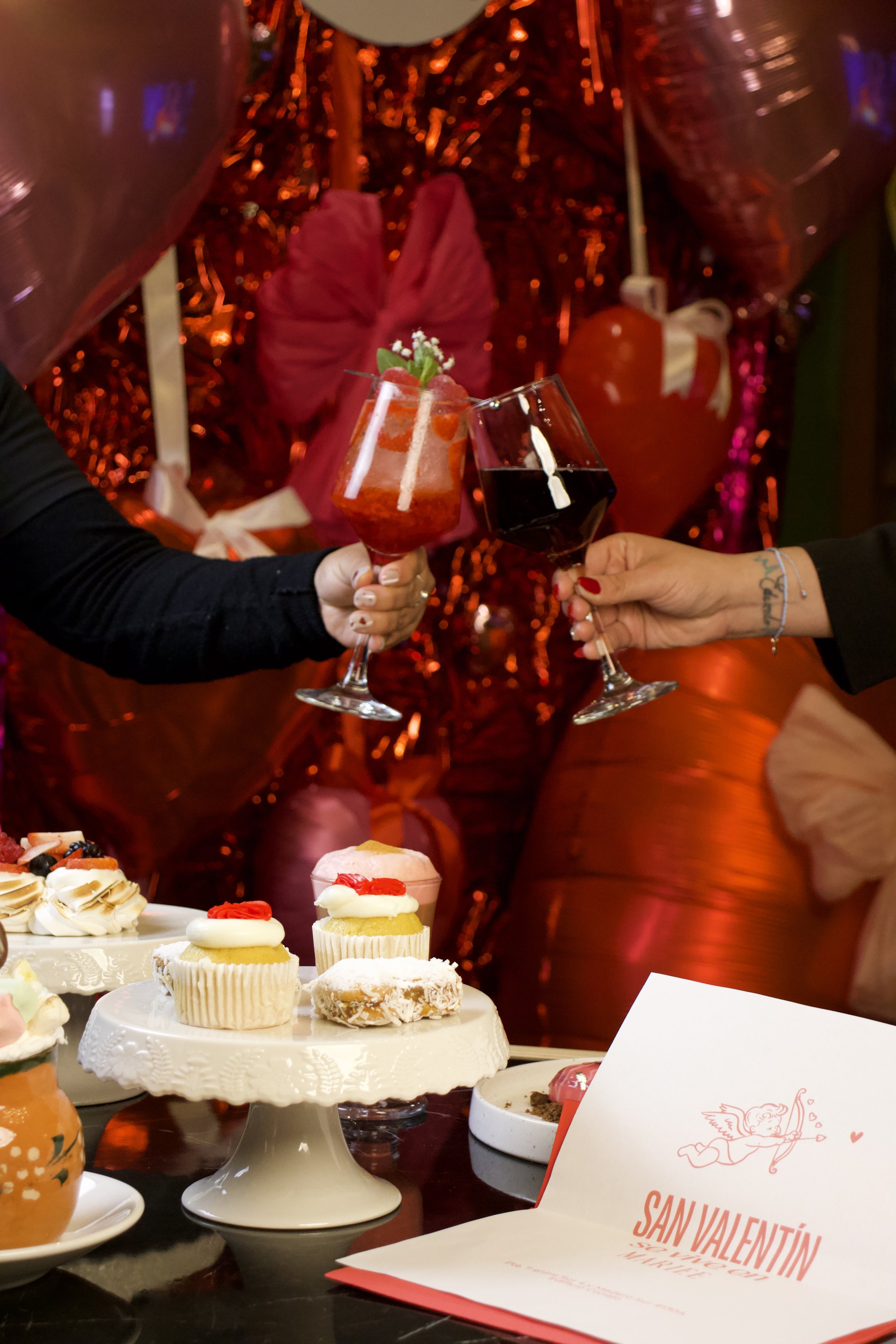 Dos personas brindando con vino tinto y bebida de color rojo en una celebración de San Valentín, con dulces y decoraciones románticas en la mesa.
