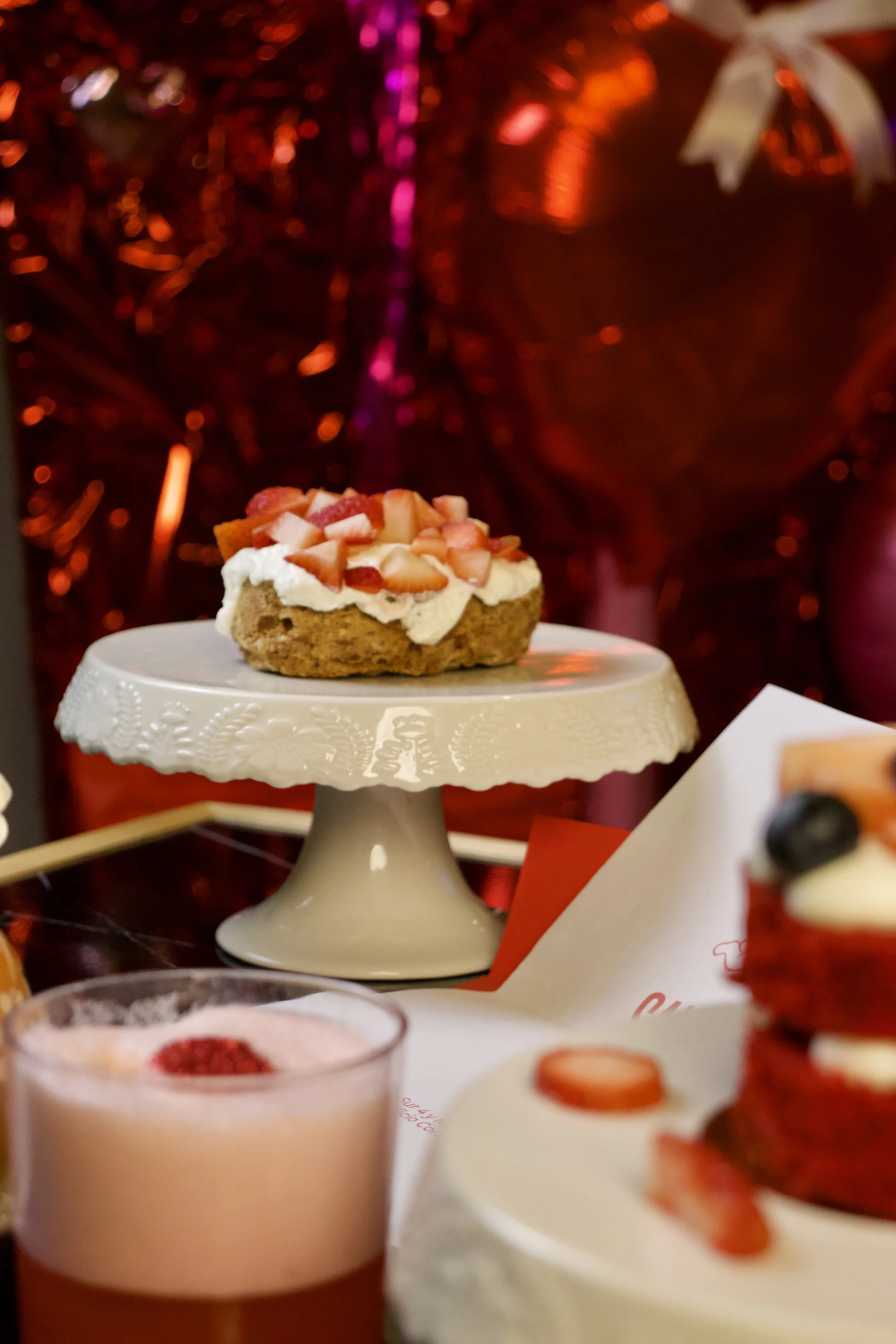 Pastel de fresa con crema y fresas en un soporte decorativo blanco y una bebida en primer plano en un ambiente festivo con decoraciones rojas.