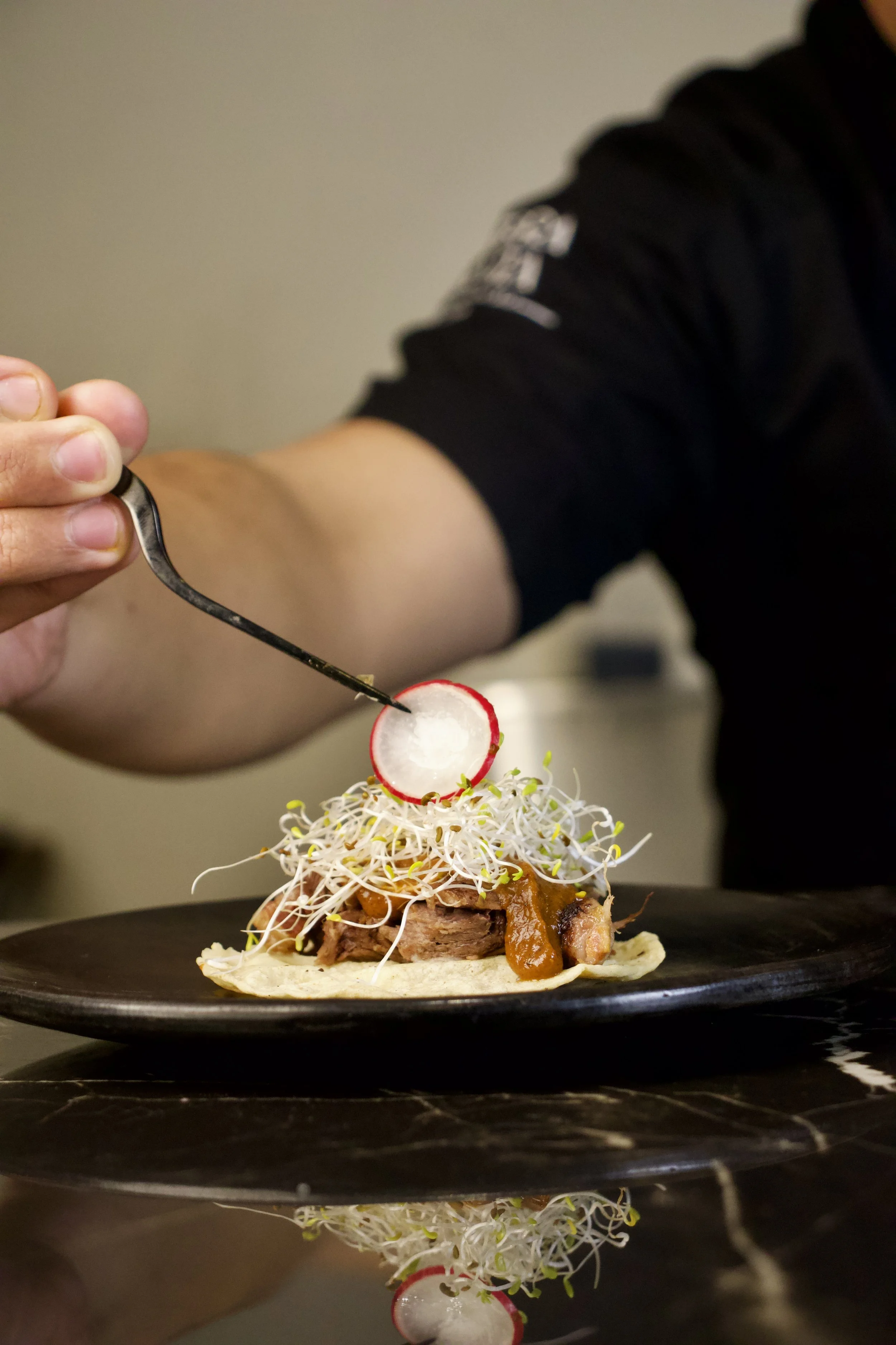 Persona decorando un plato de comida con rábanos y brotes de germinados en un plato negro.
