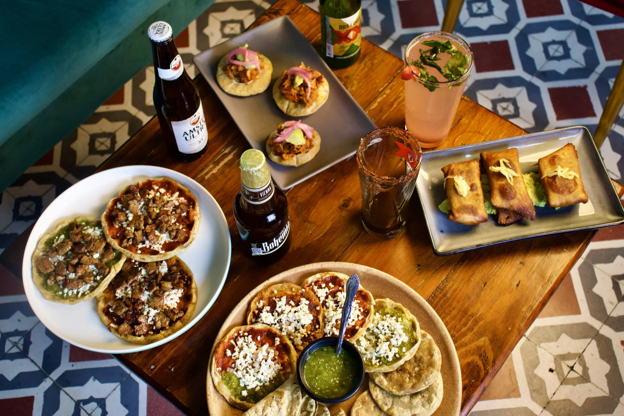 Mesa de madera con variedad de comida mexicana, incluyendo tacos, chiles en nogada, bebidas, y ingredientes diversos, sobre un piso de azulejos decorativos.