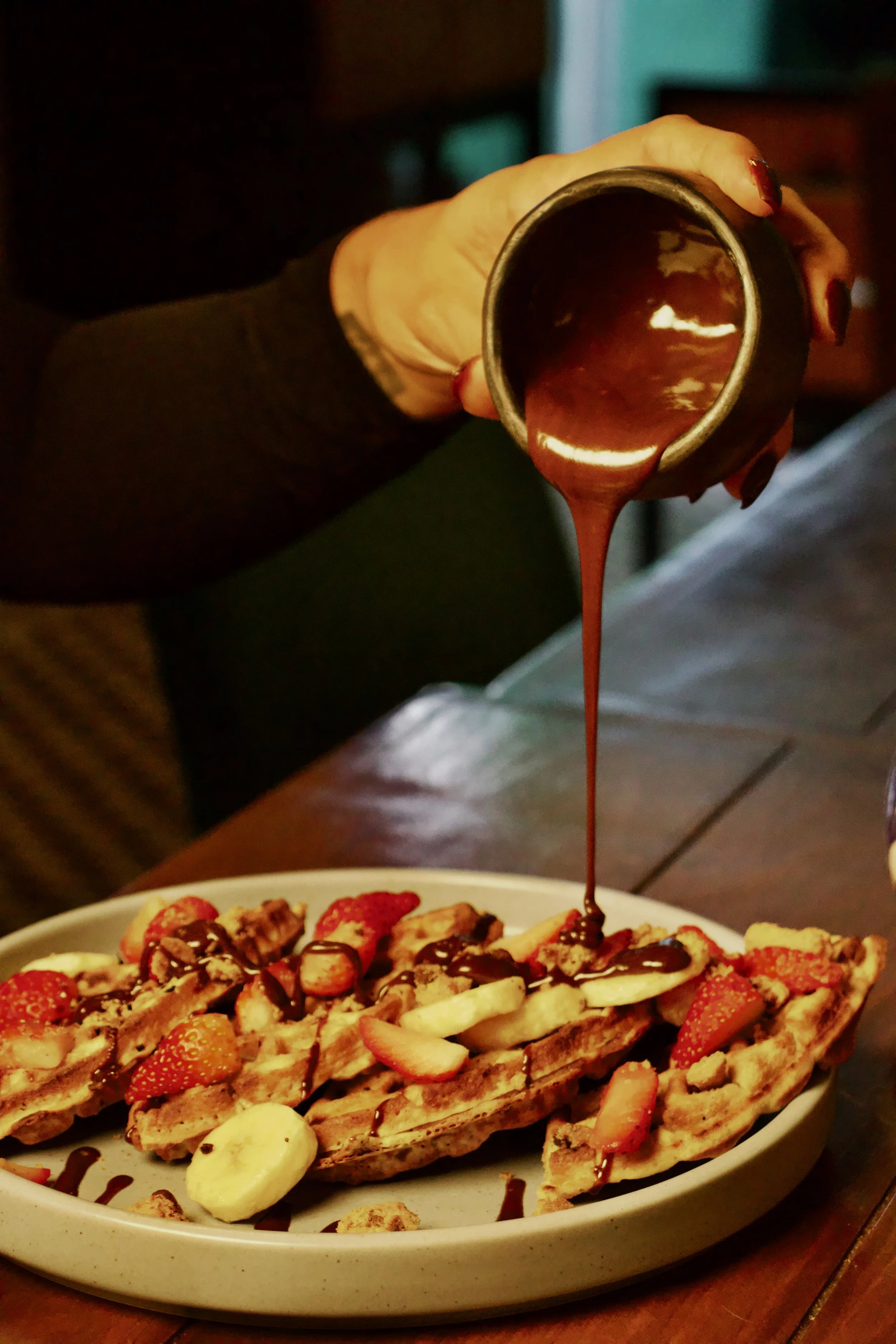 Una persona vierte salsa de chocolate sobre un plato de waffles con fruta fresca, incluyendo plátanos y fresas.