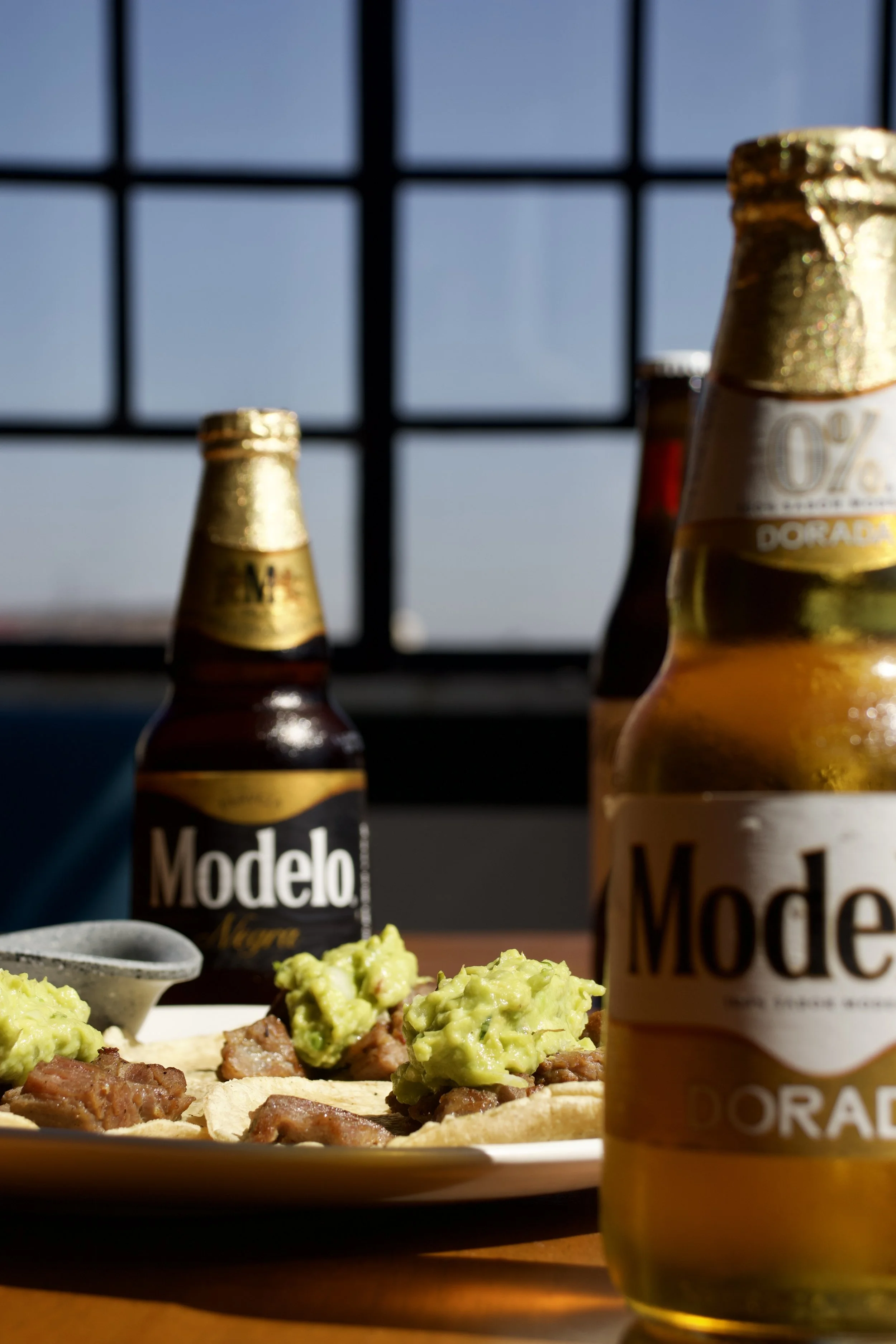 Vasos de cerveza Marca Modelo en una mesa, con una orden de comida que parece ser tacos con guacamole, en un entorno con ventana que muestra un cielo despejado.