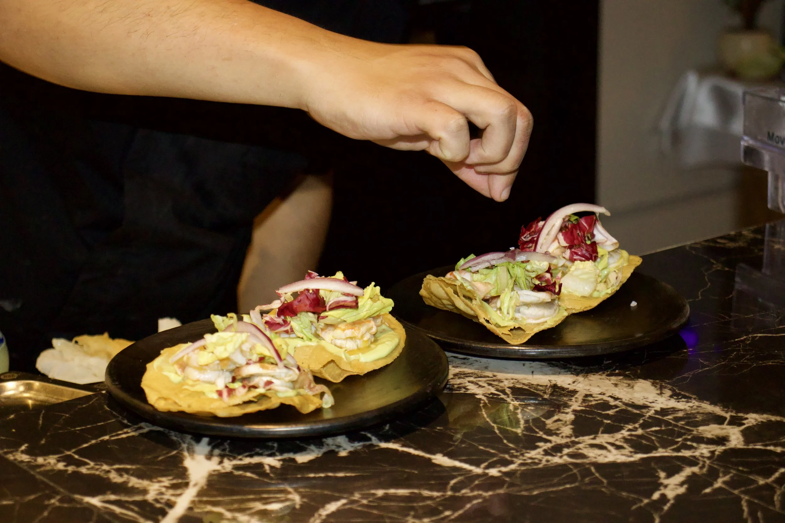 Persona colocando ingredientes en tacos de tortilla con plátano, ensalada y mariscos en un plato negro