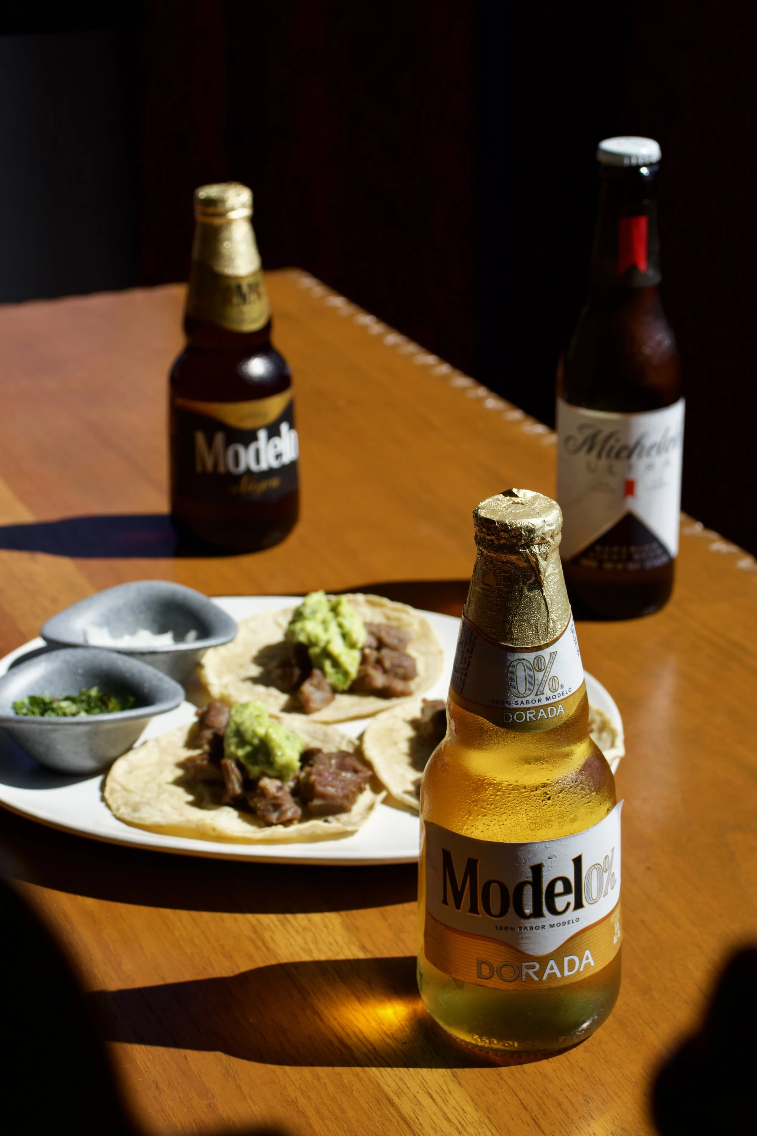 Tres botellas de cerveza Modelo Dorada y una botella de Michelob en una mesa de madera, con un plato de tacos con carne, guacamole y salsas.