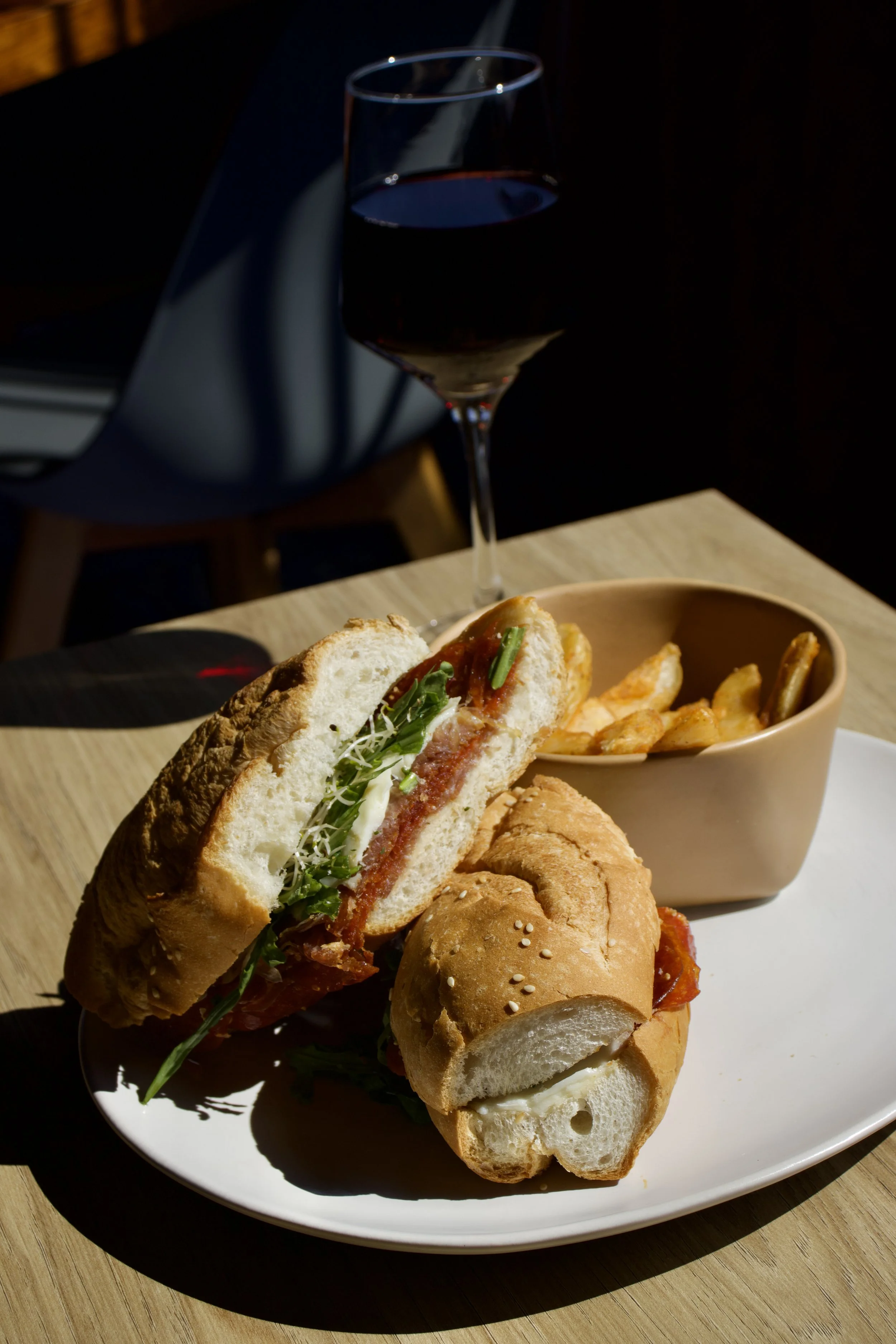 Un sándwich en pan de baguette con lechuga, tocino y salsa acompañado de papas fritas en un plato, con una copa de vino tinto al fondo.