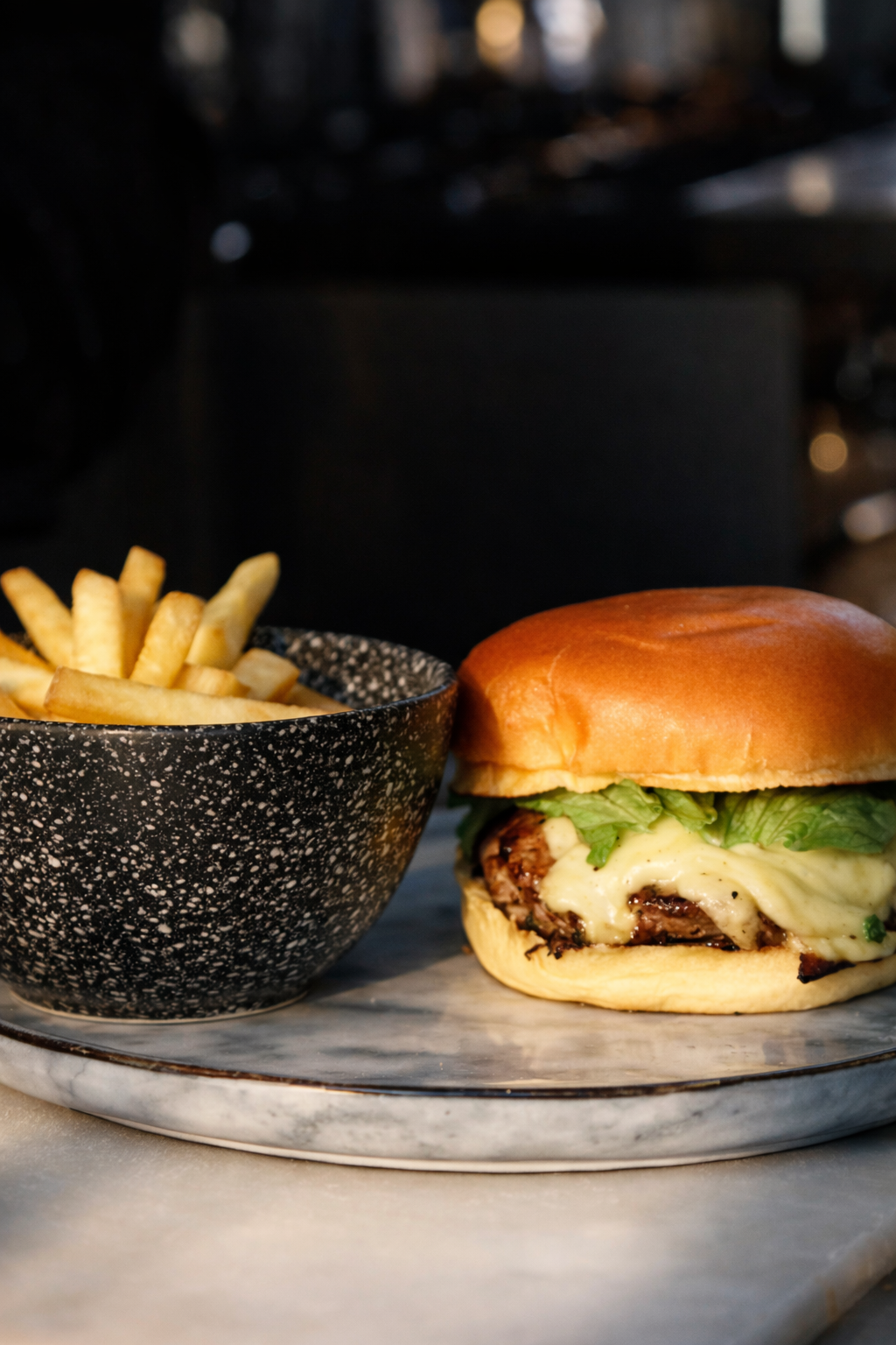 Una hamburguesa con pan dorado, lechuga, queso y carne, acompañada de un bol de papas fritas en un plato de mármol.
