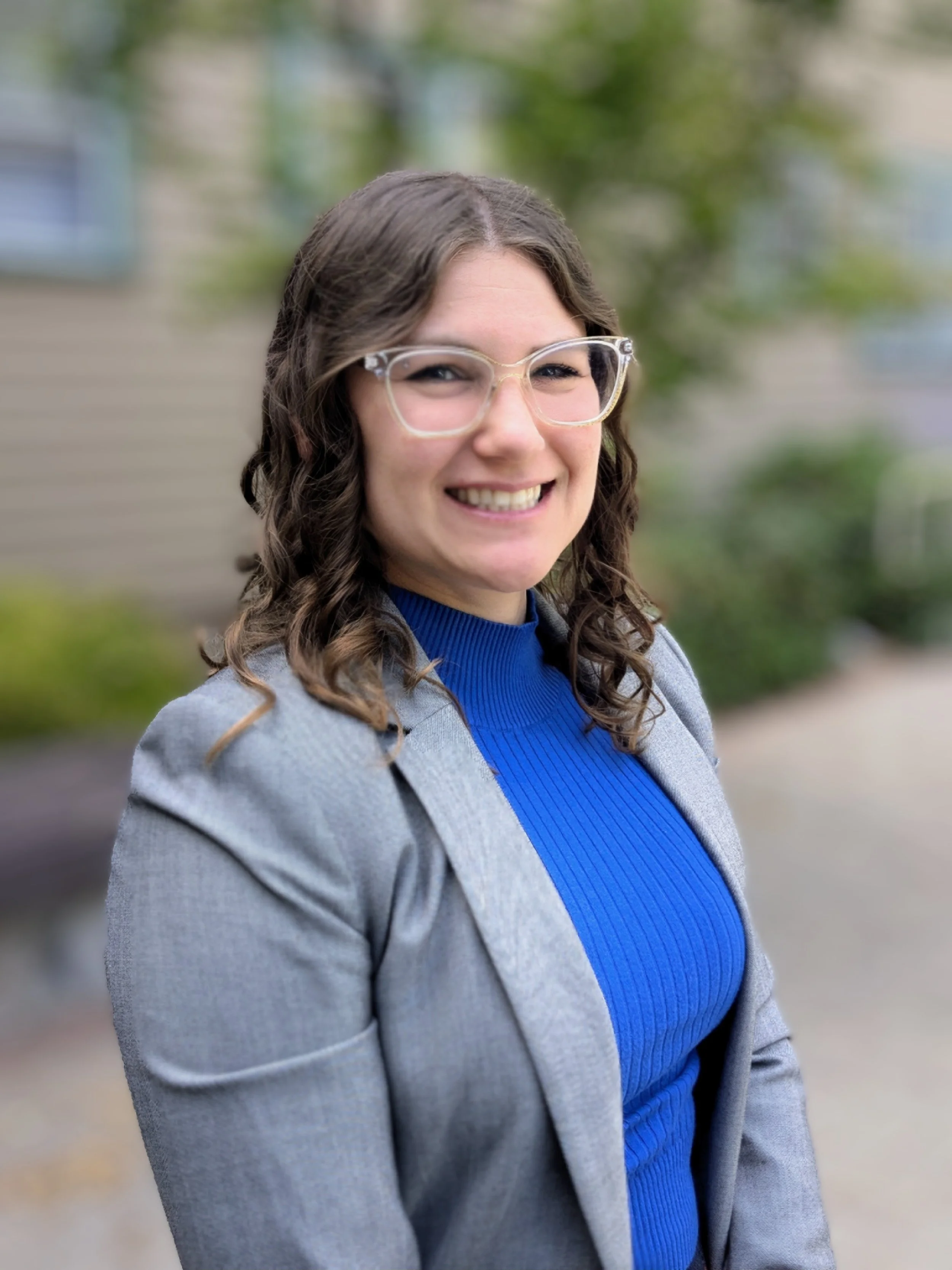 A woman with glasses and curly brown hair smiling outdoors, wearing a gray blazer and bright blue turtleneck sweater.