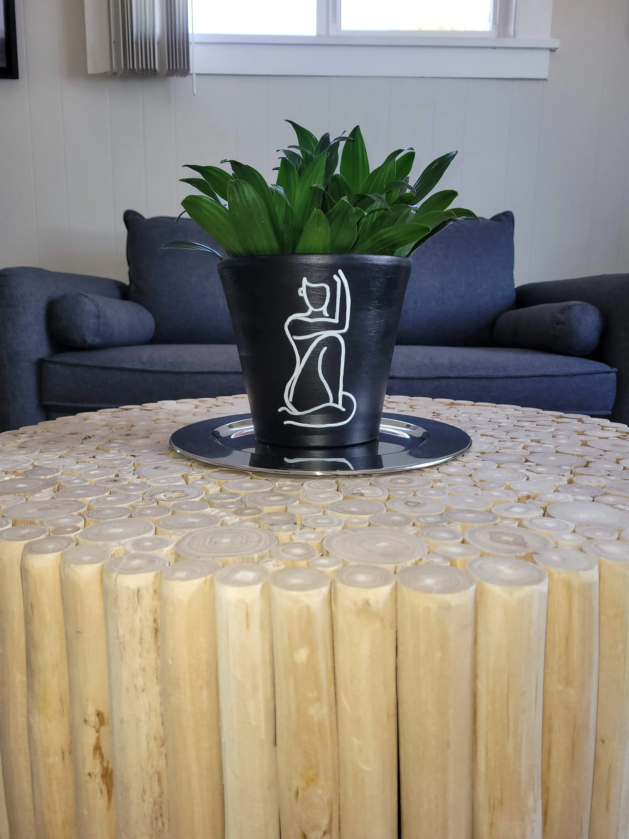 A black flower pot with a white line drawing of a kneeling woman, sitting on a table made of wood slices, containing green leafy plants, with a silver tray underneath, in a living room with blue couch and white wall with window.
