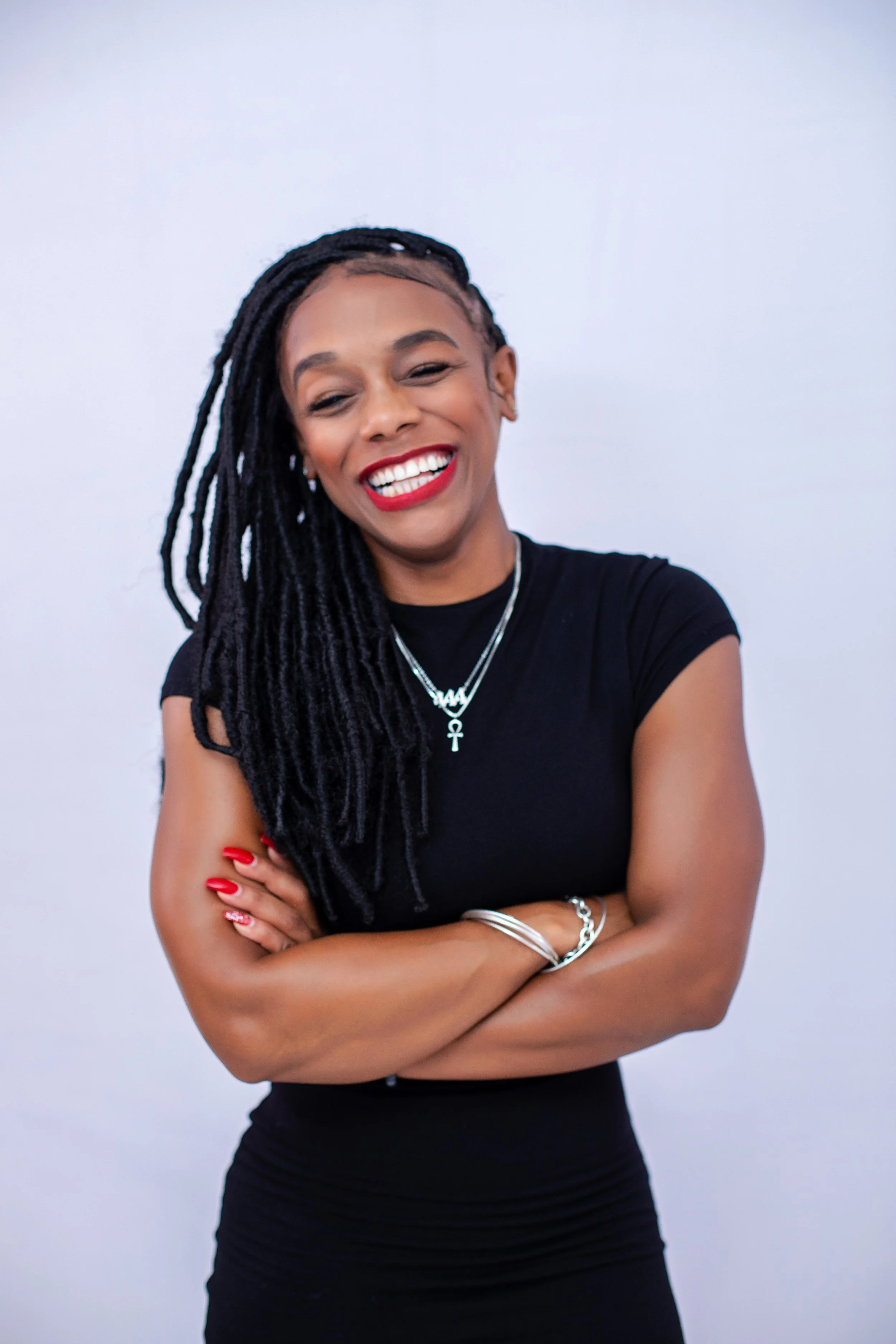 A woman with long dreadlocks, wearing a black shirt, smiling with arms crossed, against a plain white background.