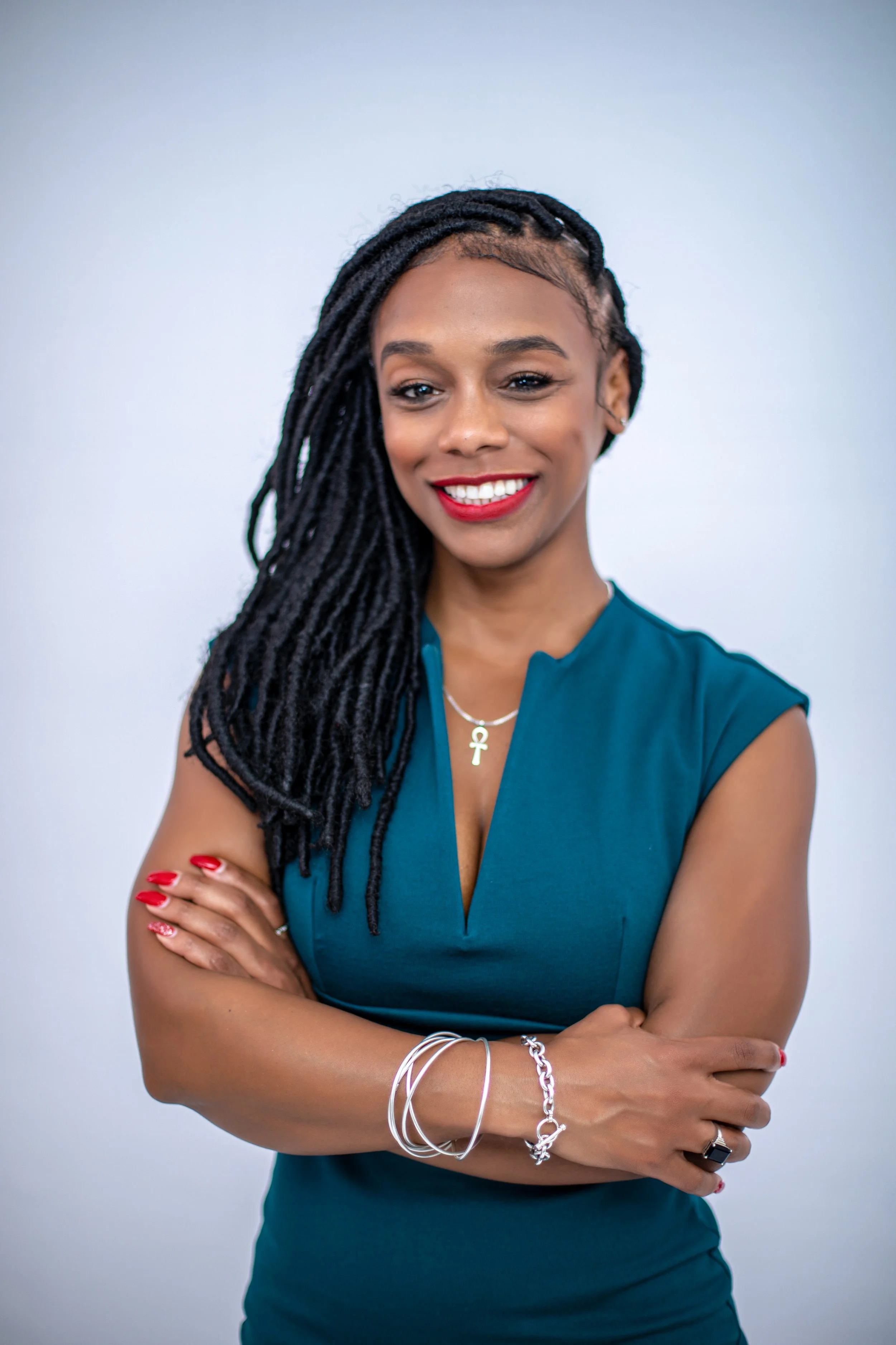 Woman with long dreadlocks wearing a teal sleeveless top, smiling, with arms crossed, against a plain white background.