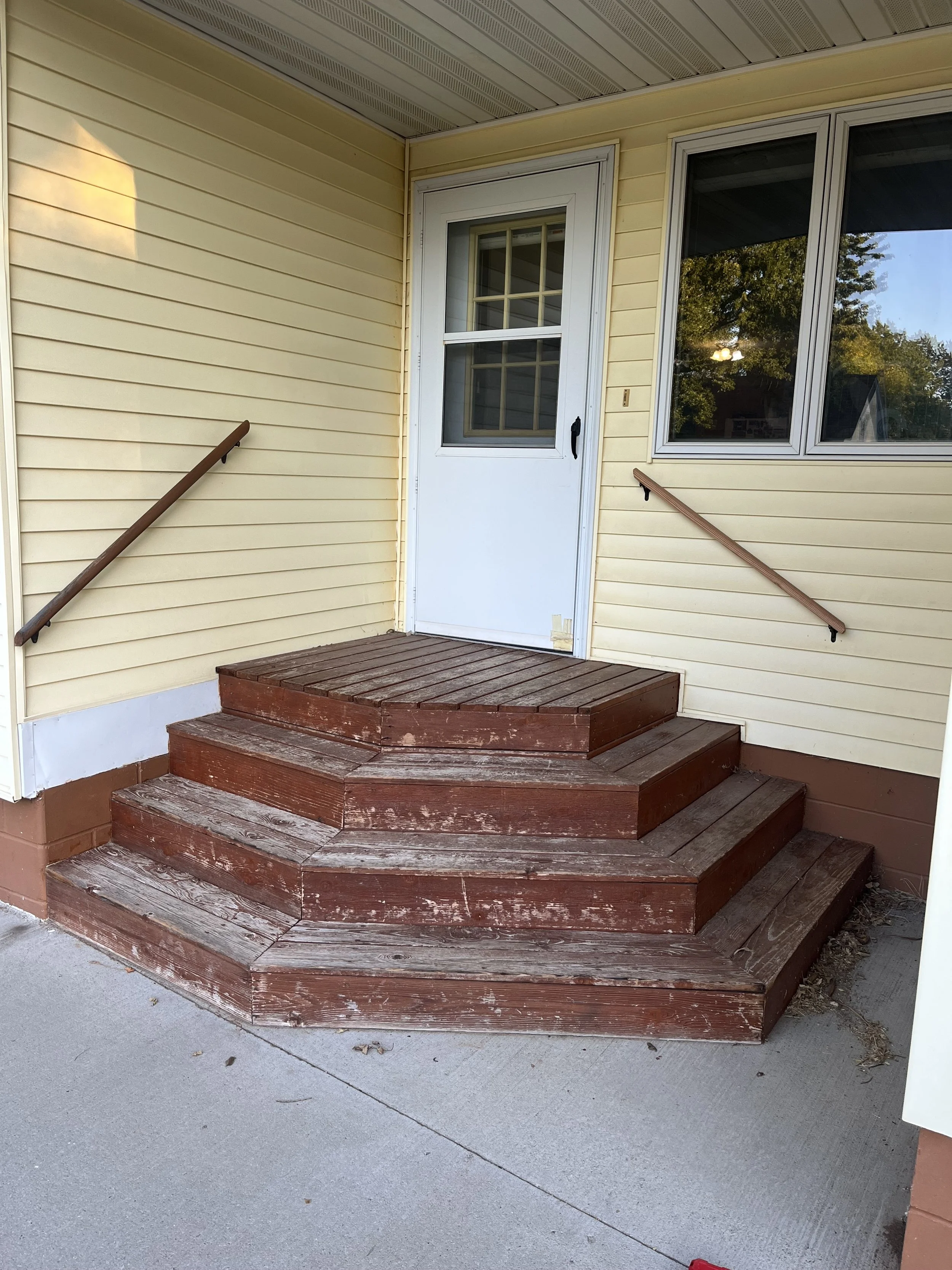 Exterior front porch with wooden stairs, yellow siding, white door with glass panels, and a window reflecting outdoor trees.