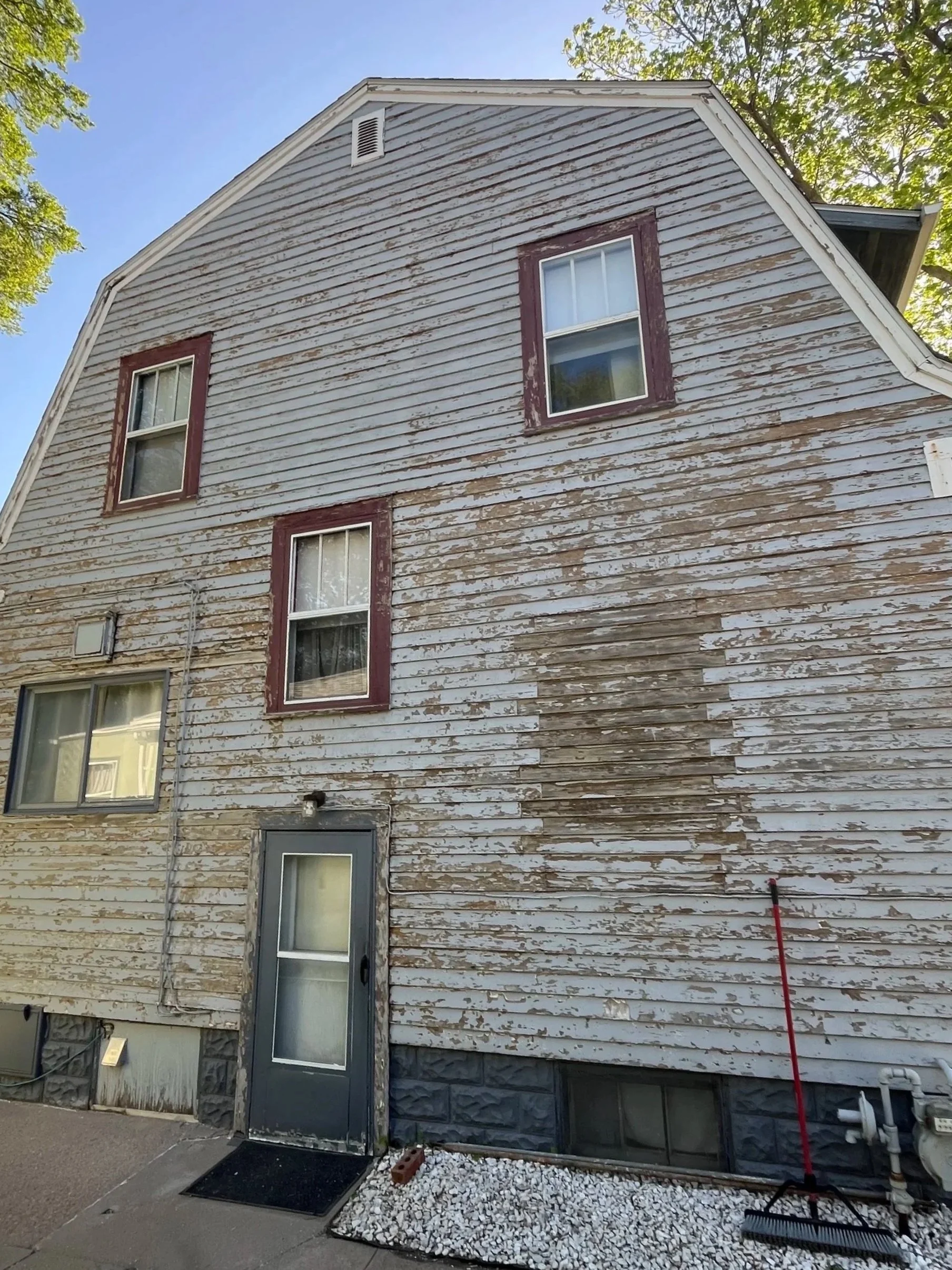 A three-story house with peeling white paint on wooden siding, two windows on the second floor, and a door on the ground floor. The house has a pitched roof and is surrounded by trees and sunlight.