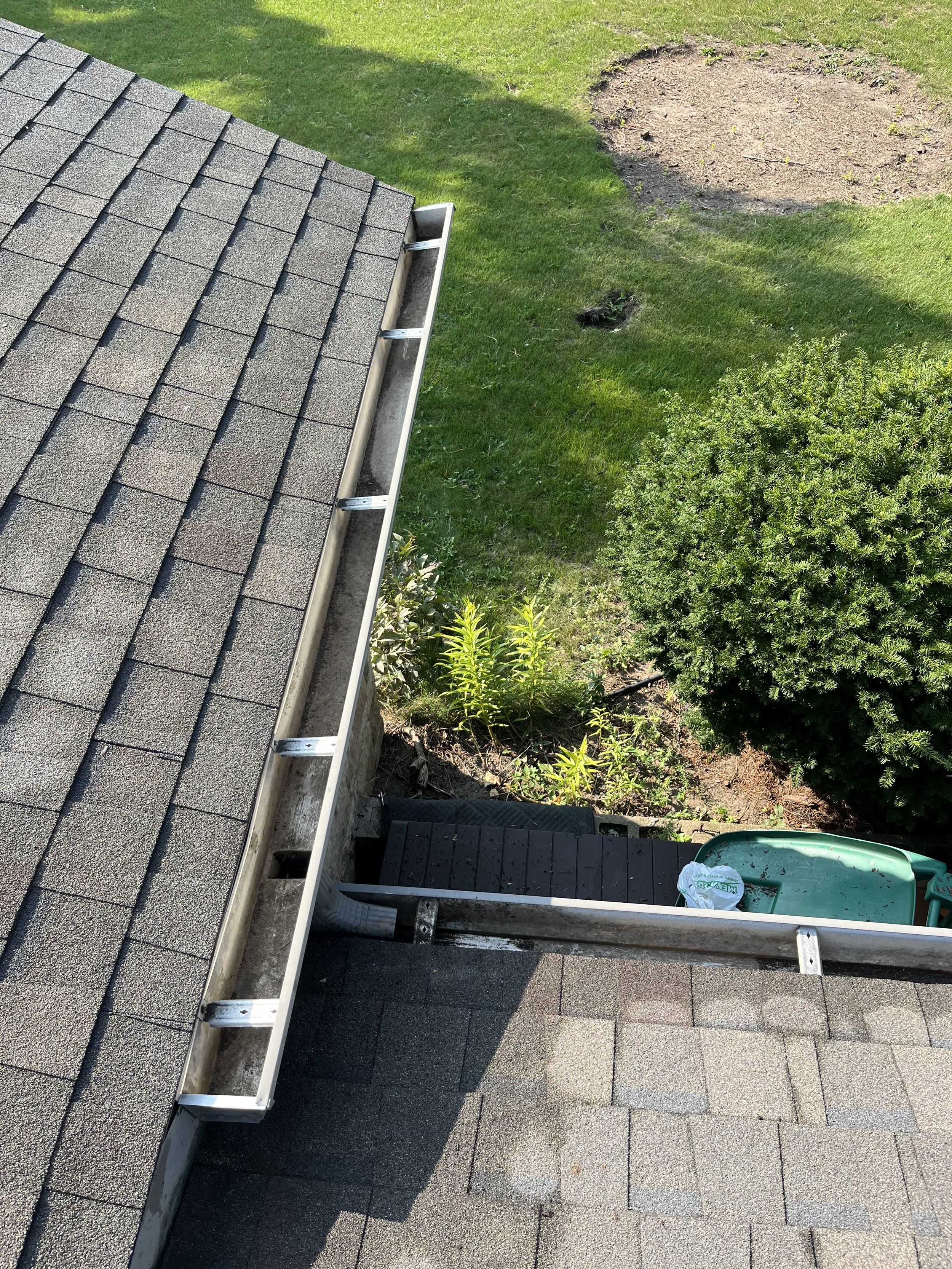 View of house roof with shingles, gutter, and a ladder structure, overlooking a garden with grass and bushes.