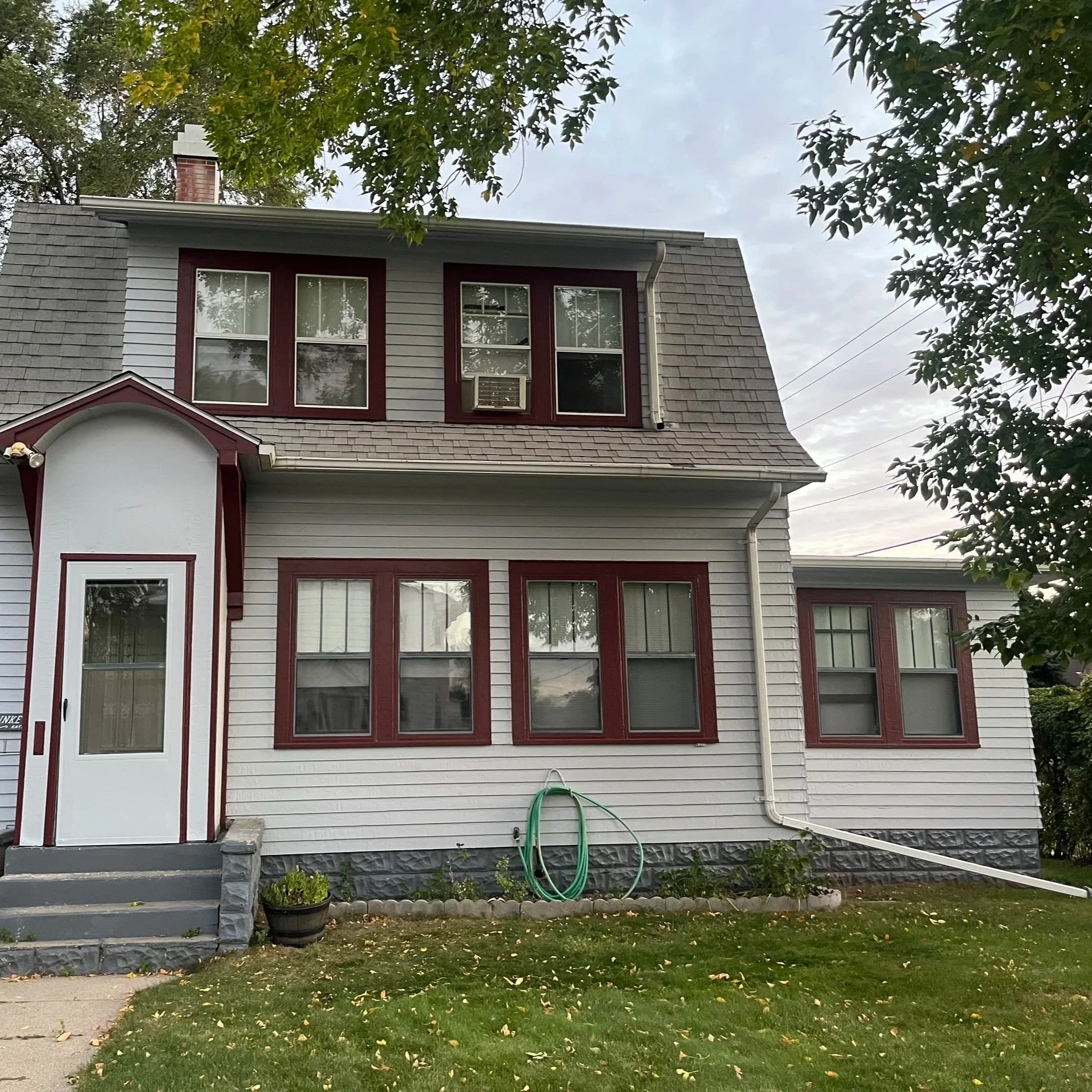 Front view of a two-story house with white siding, maroon window frames, and a stone foundation. There is a small set of steps leading to the front door, which is white with a window, and a potted plant on the left side of the steps. Green garden hose is coiled on the grass in front of the house. Leafy trees partially frame the house at the top and right side.