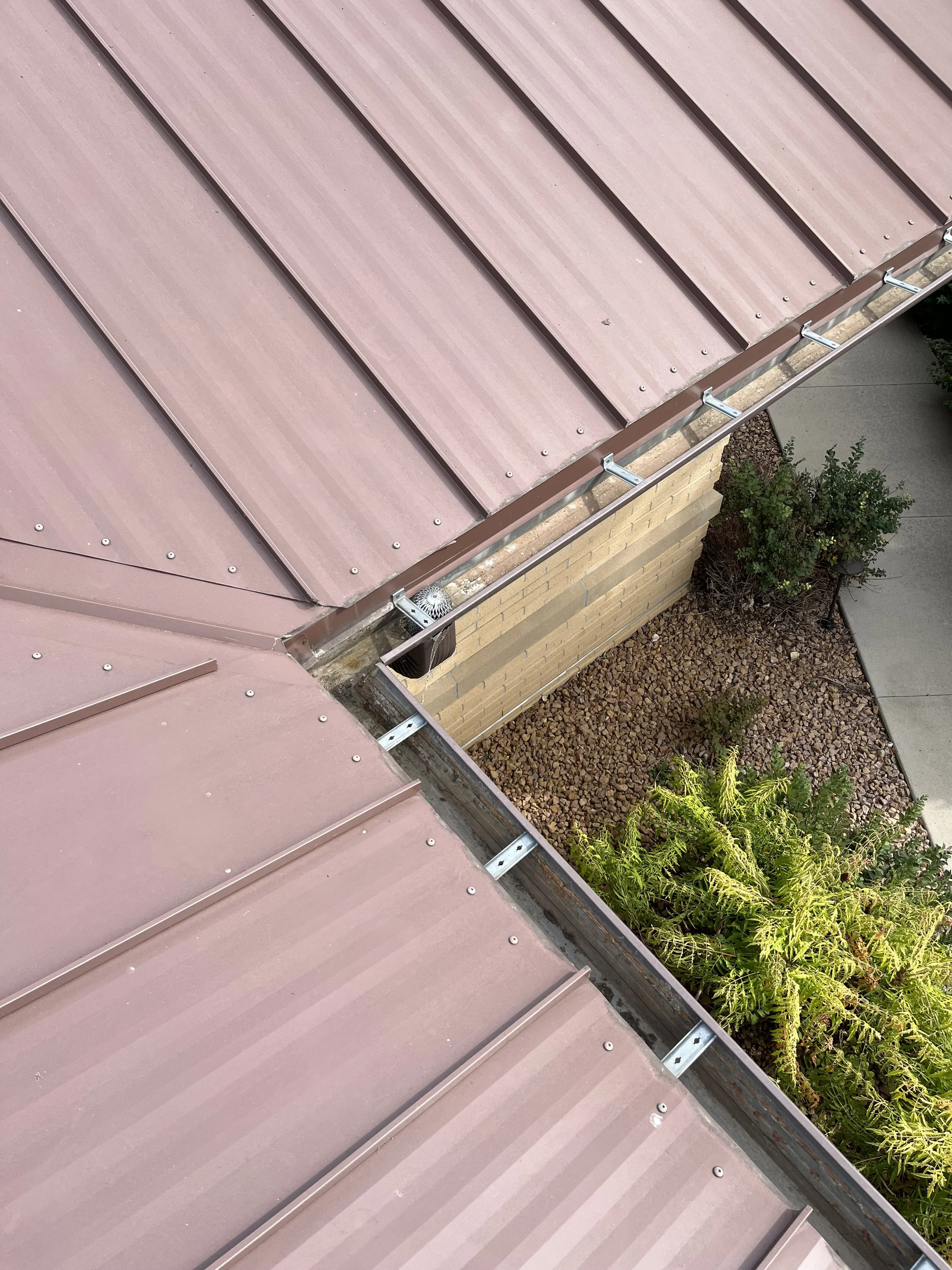 Aerial view of a metal roof with visible screws and a gutter, next to a brick wall and landscaped area with plants.