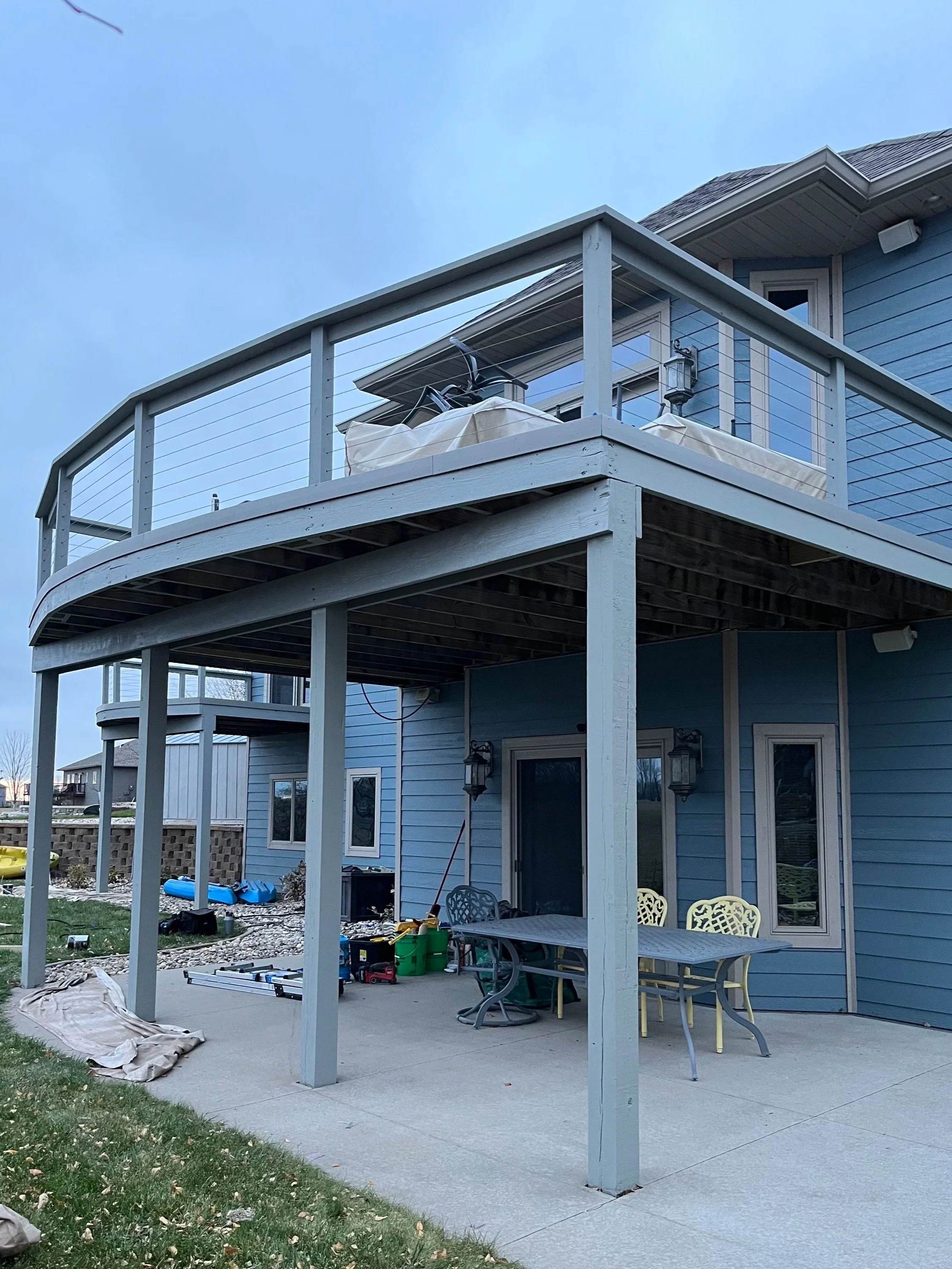 Backyard patio of a blue house with an upper and lower deck, outdoor furniture, and various tools and equipment.