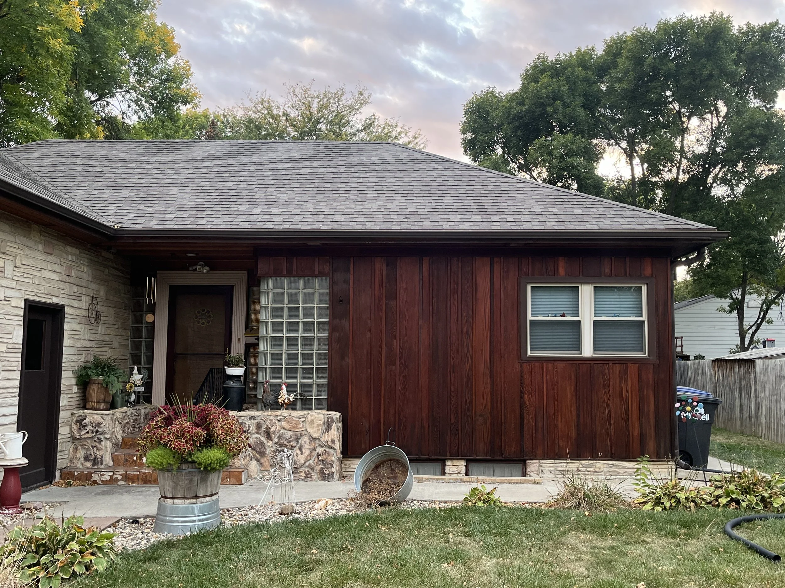 A house with a stone and wooden exterior, a sloped shingled roof, green lawn, potted plants, and some yard decorations. There are trees and a cloudy sky in the background.