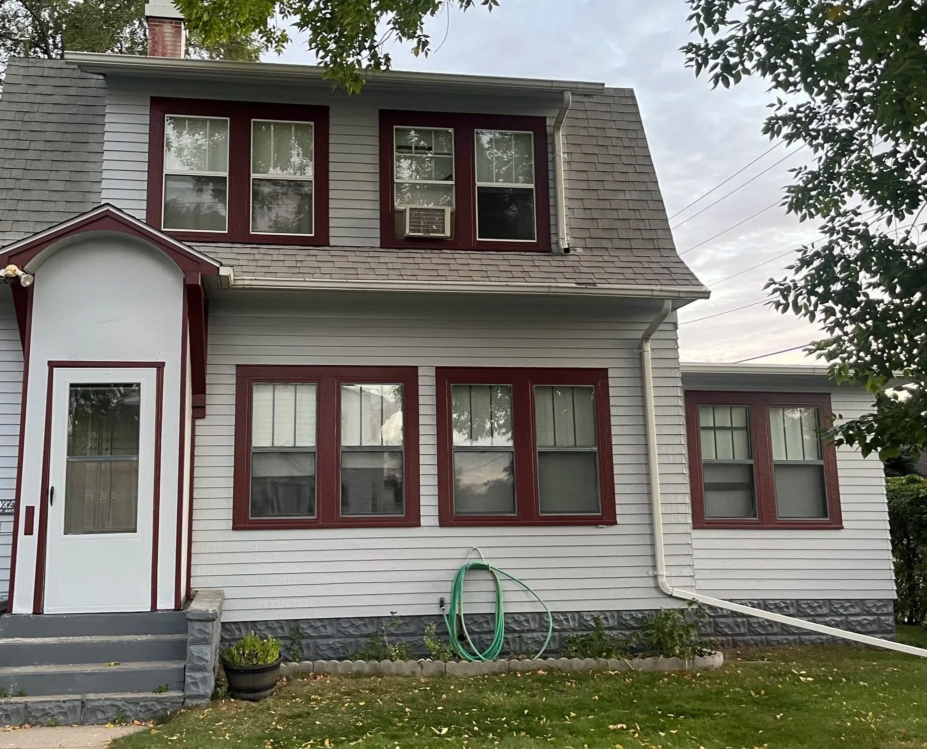 A two-story house with gray siding, red trim around the windows and door, a small front porch with gray steps, and a green garden hose coiled against the house at ground level. There are trees and a cloudy sky in the background.
