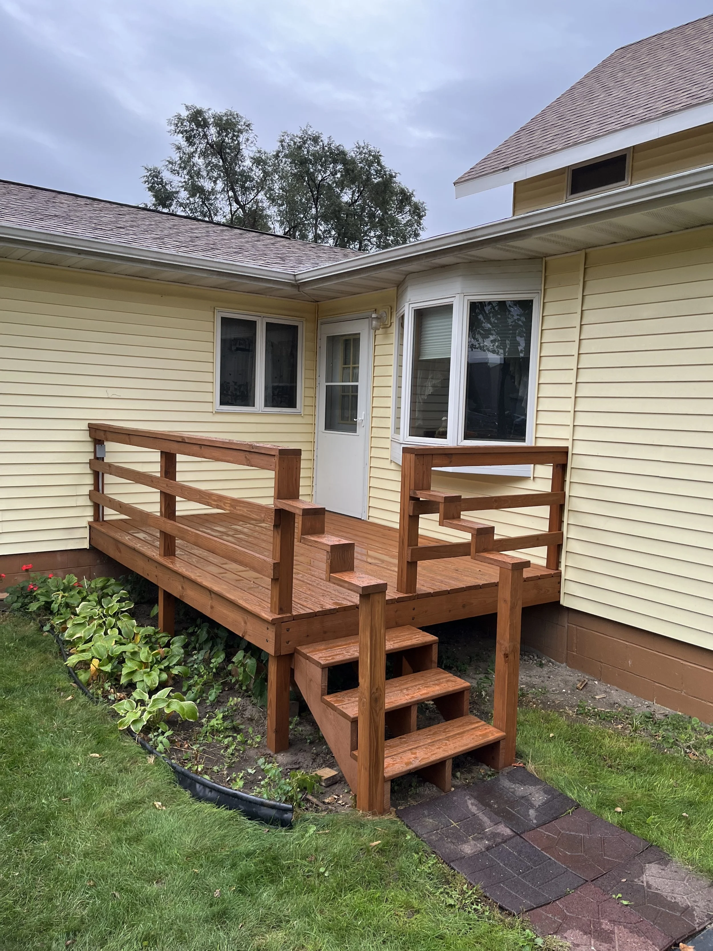 Front porch with newly built wooden ramp and stairs leading to a white door on a yellow house, with a garden bed and green grass in the yard.