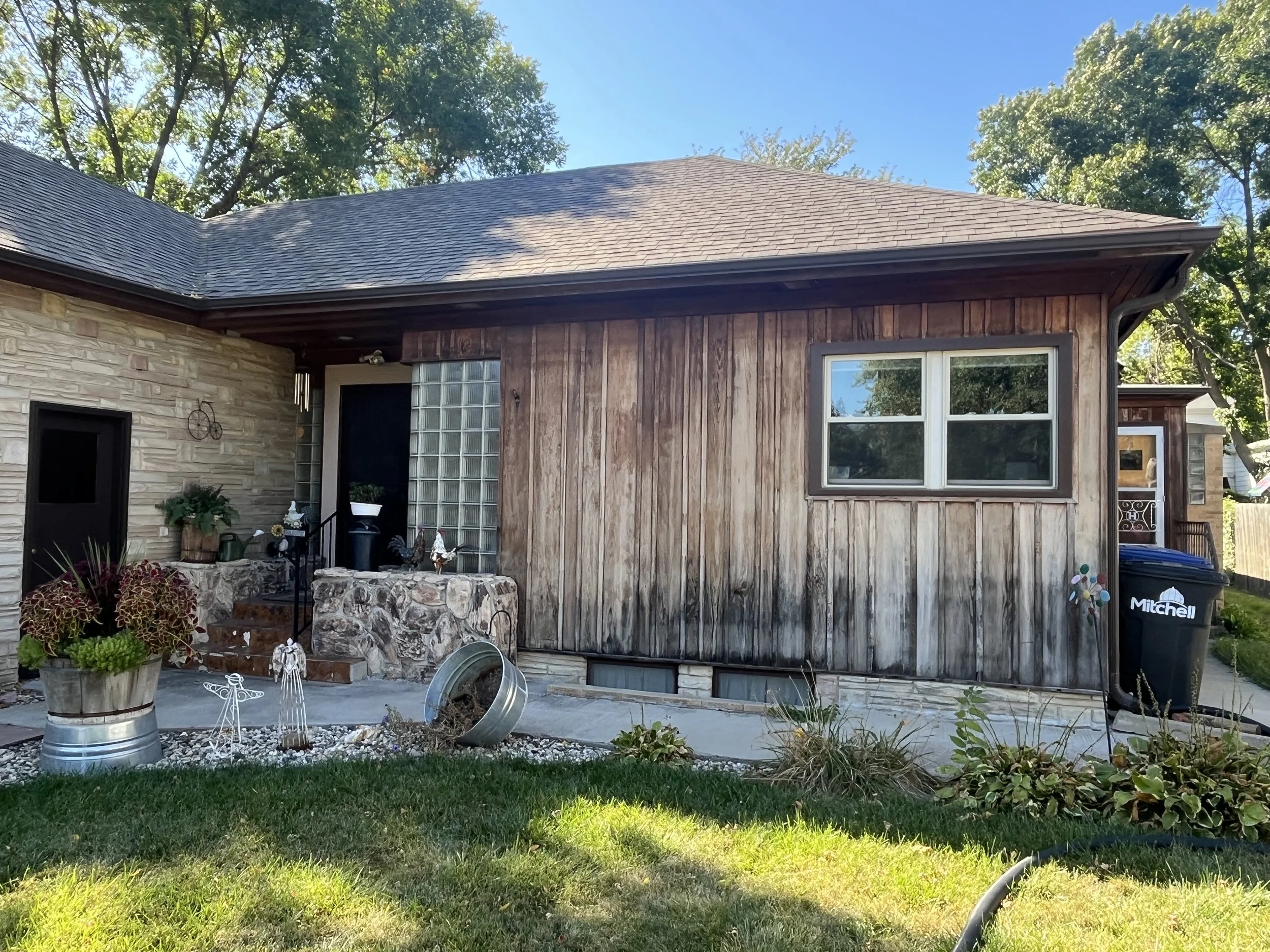 Backyard of a house with a wooden and brick exterior, two windows, and a small porch area decorated with potted plants and garden ornaments, with a grassy lawn in the foreground.