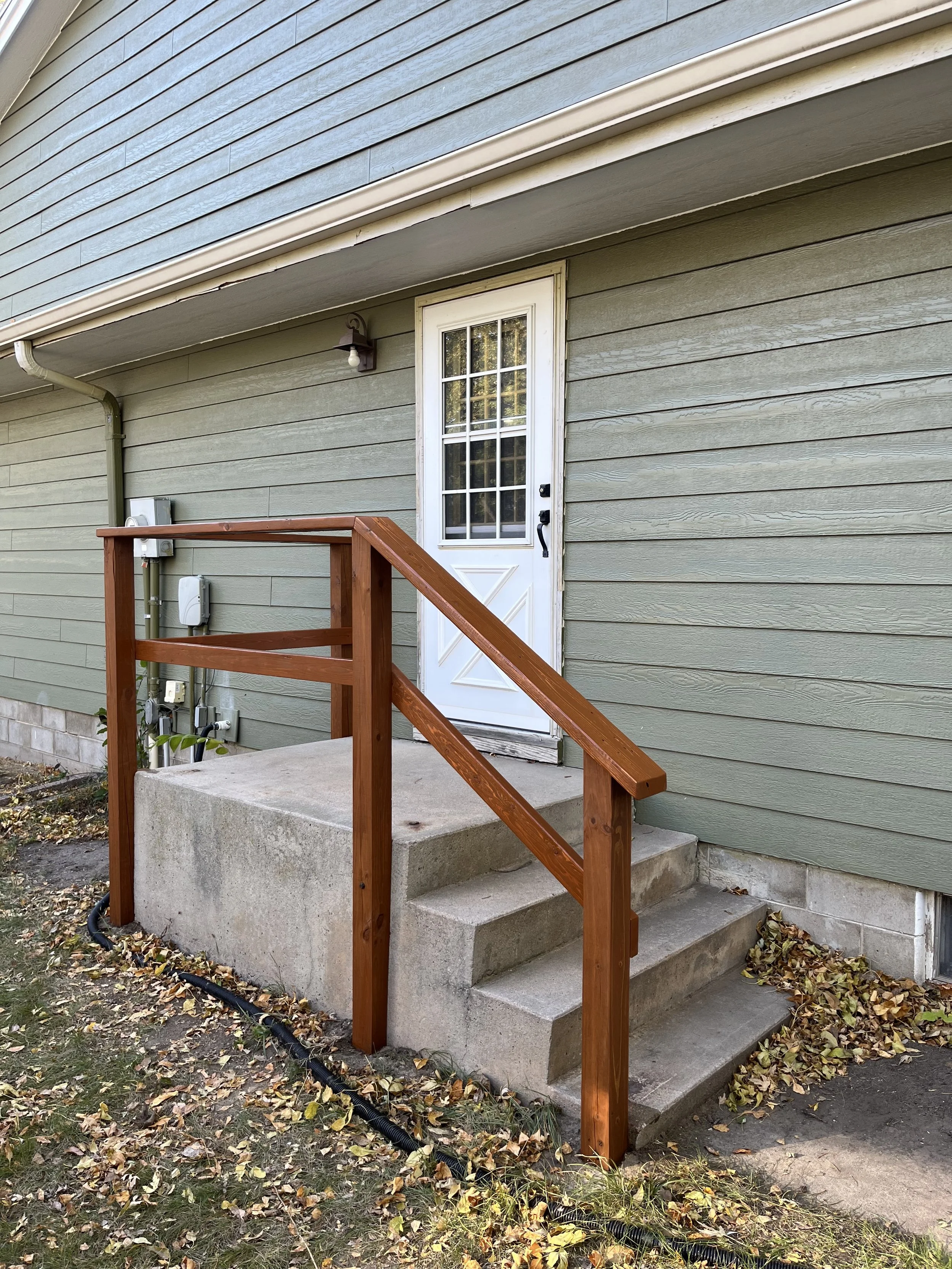 Front steps with a small concrete landing, new wooden railing, white door with glass panel, green horizontal siding, outdoor light, and some fallen leaves.