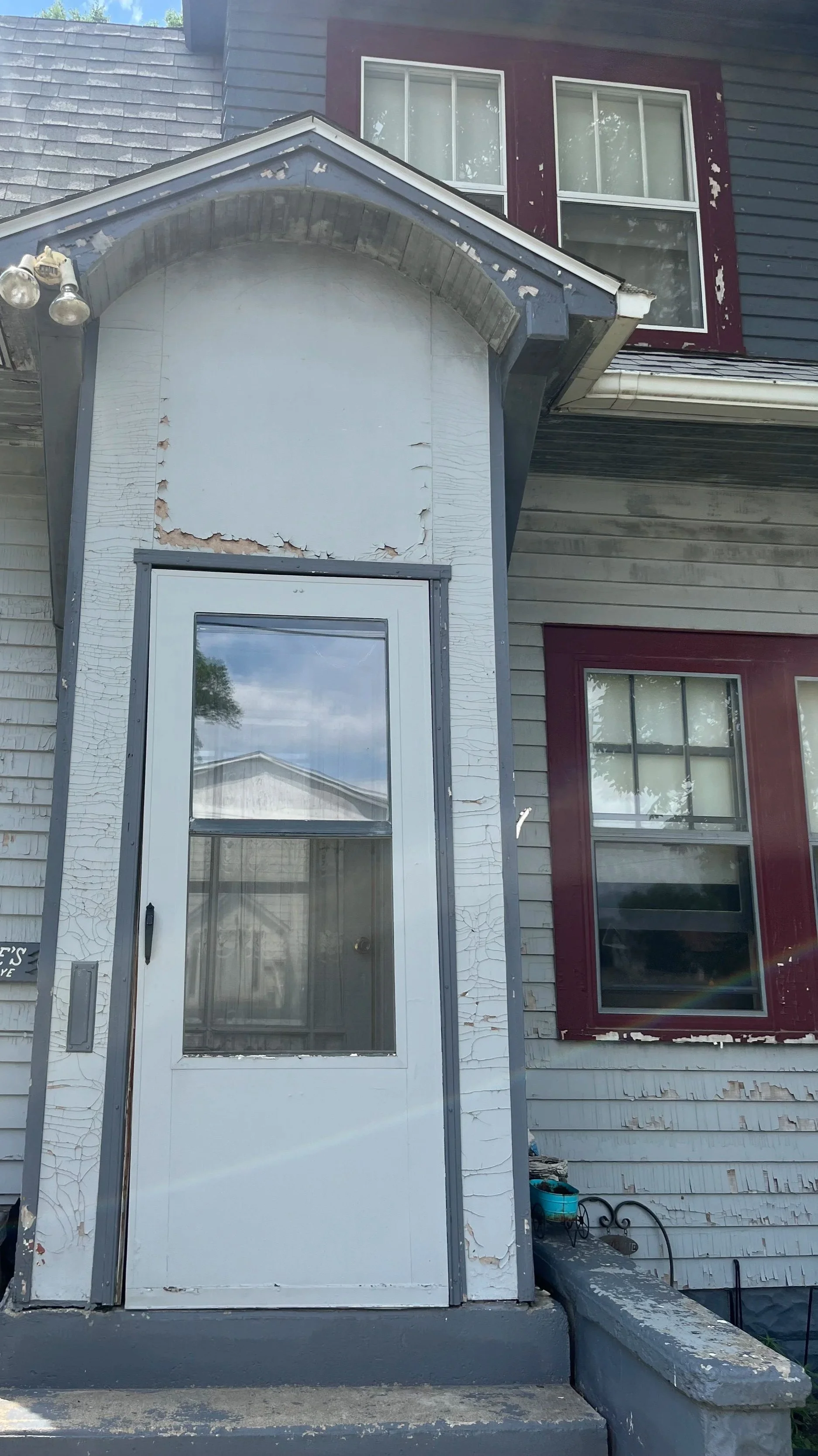 Front entrance of a house with a white door and cracked paint, peeling siding, and two windows with maroon trim.