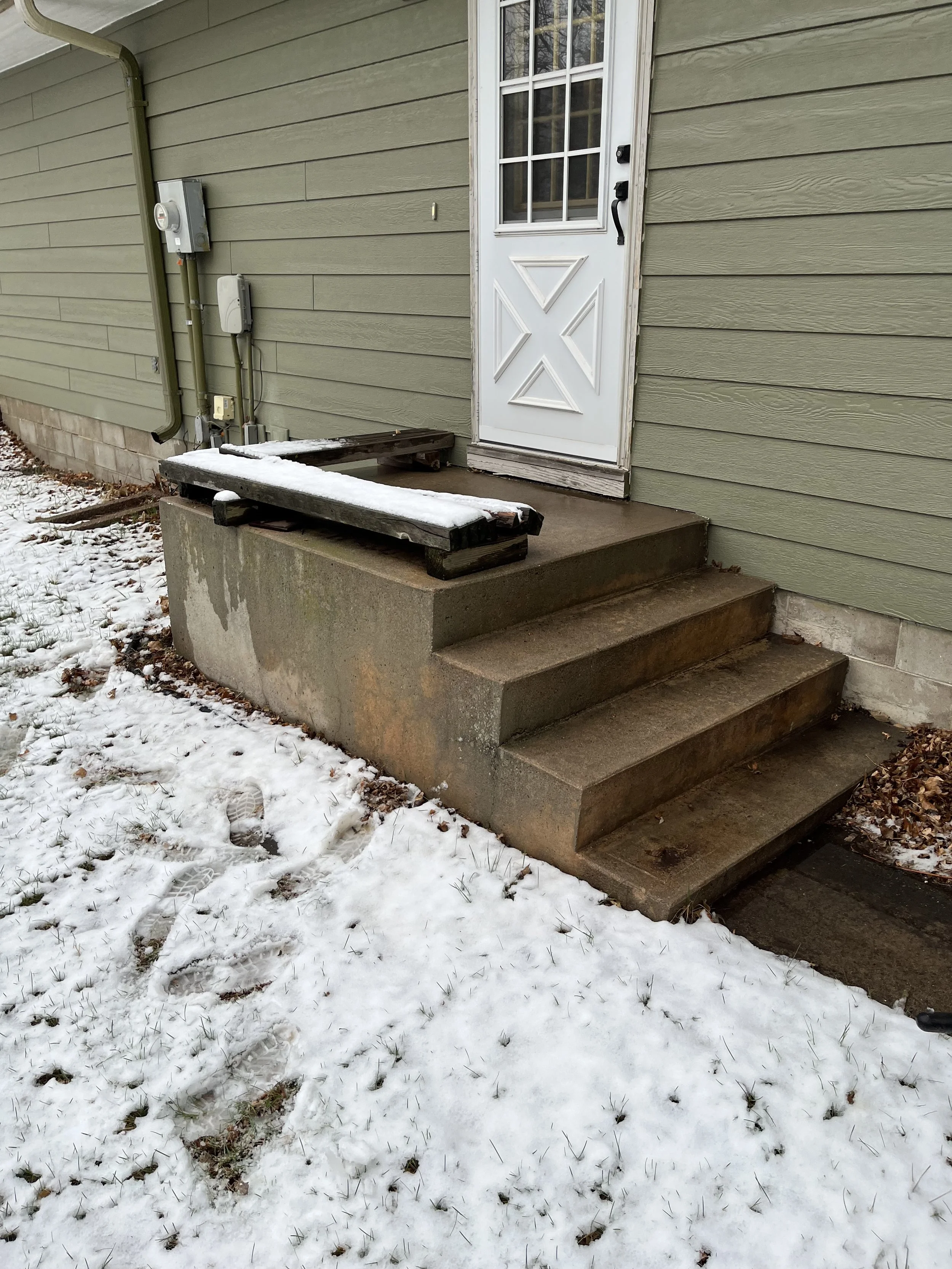 Concrete steps leading to a white door with a window on a house with green siding. Snow on the ground and on a wooden pallet on the small concrete landing.