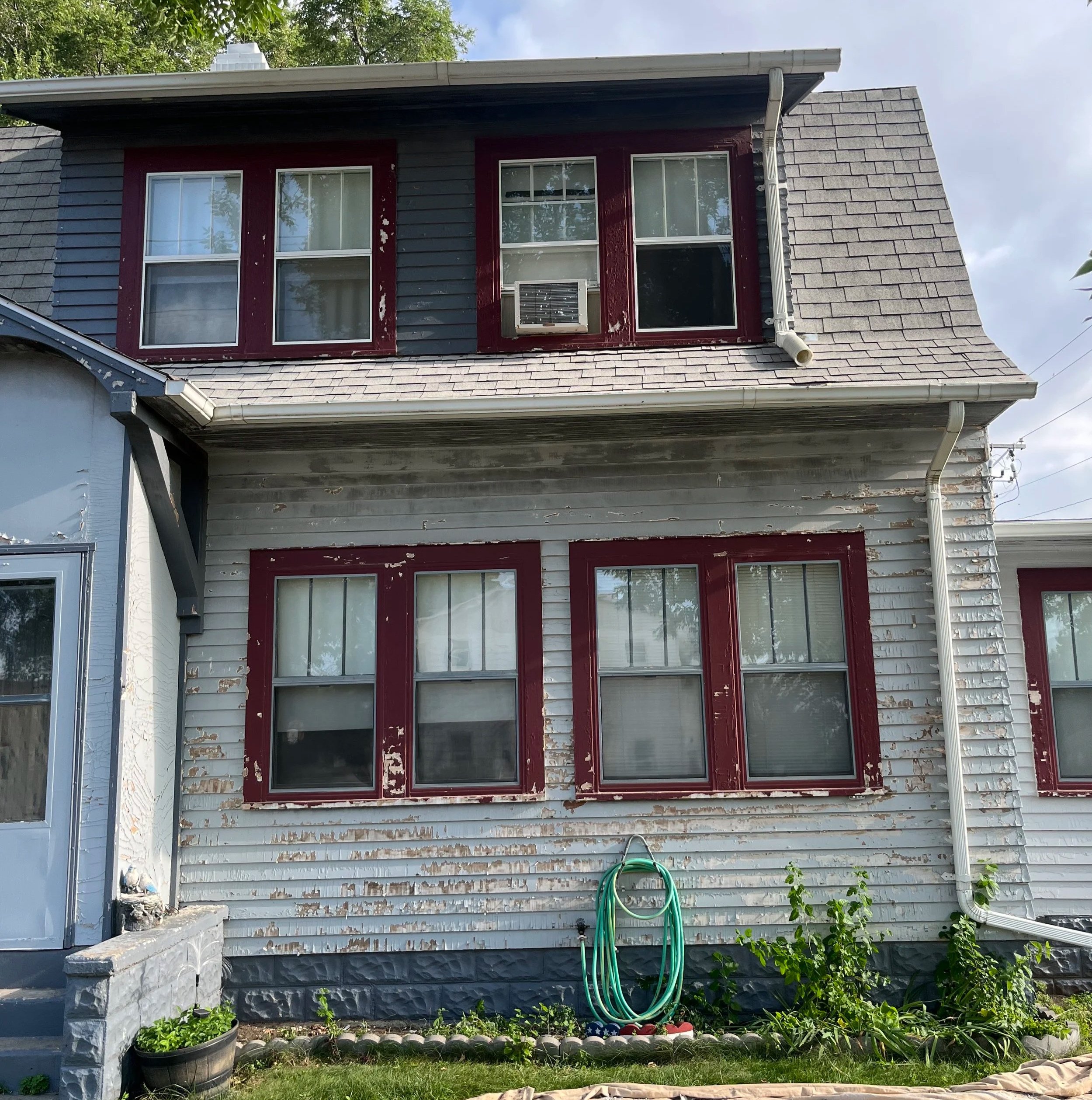 Front exterior of a two-story house with weathered gray siding and red window frames, some peeling paint, a garden hose coiled on the ground, and plants growing along the foundation.