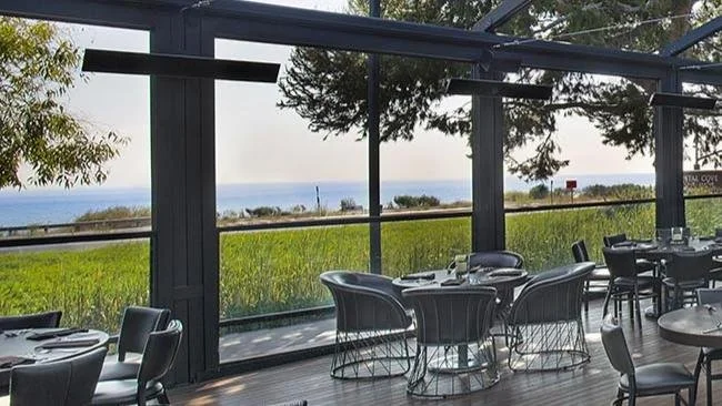 Close-up view of an elegant oceanfront restaurant patio with retractable glass walls and dark steel framing. Modern round tables and woven chairs sit atop warm wood flooring, overlooking lush greenery and the sea beyond.