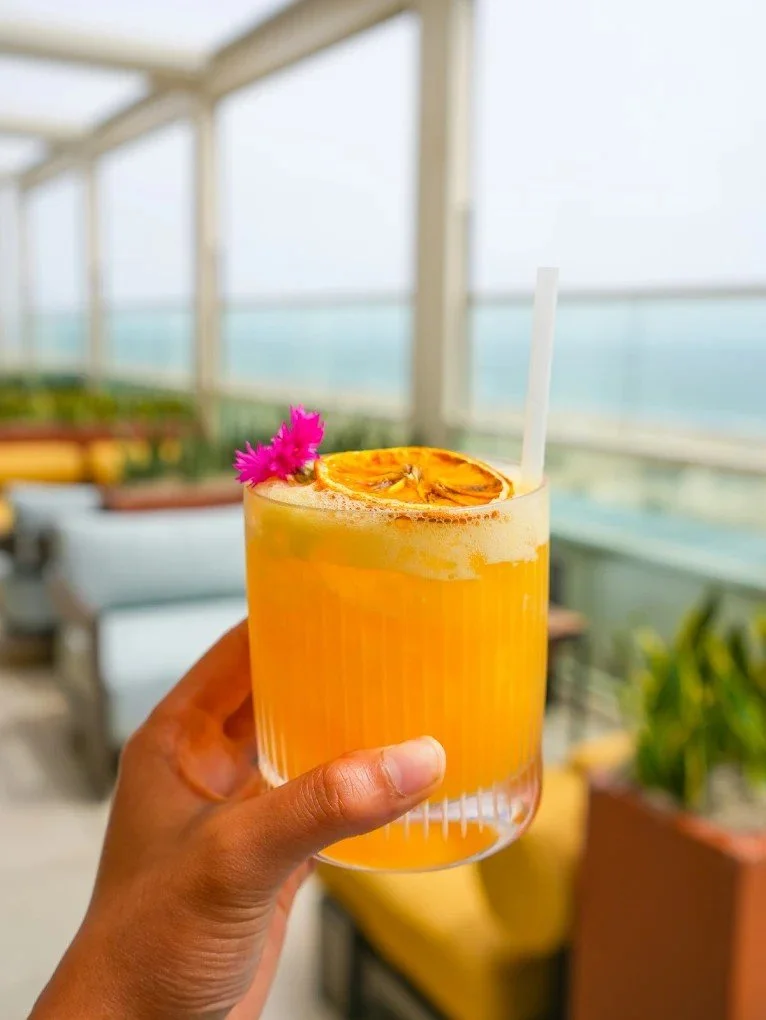Hand holding cocktail with pier and ocean view in the background behind glass walls.