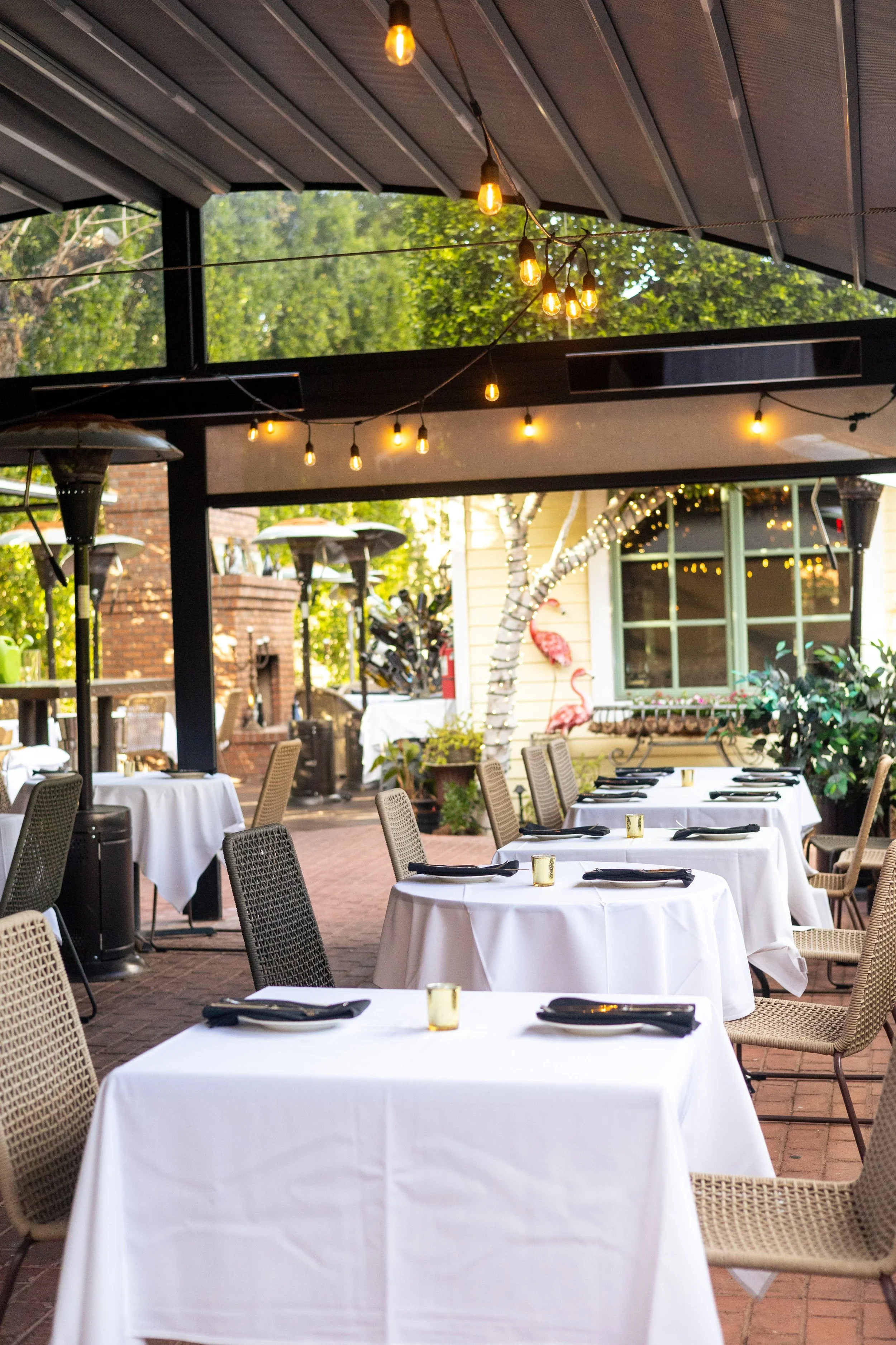House Brasserie courtyard tables beside a brick fireplace and twinkle-lit trees beneath a black aluminum canopy.