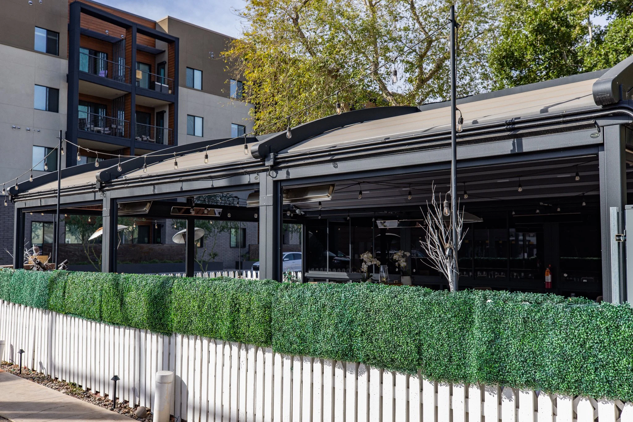 Side view of House Brasserie’s adaptive enclosure with motorized glass walls and retractable roof behind a boxwood privacy hedge.