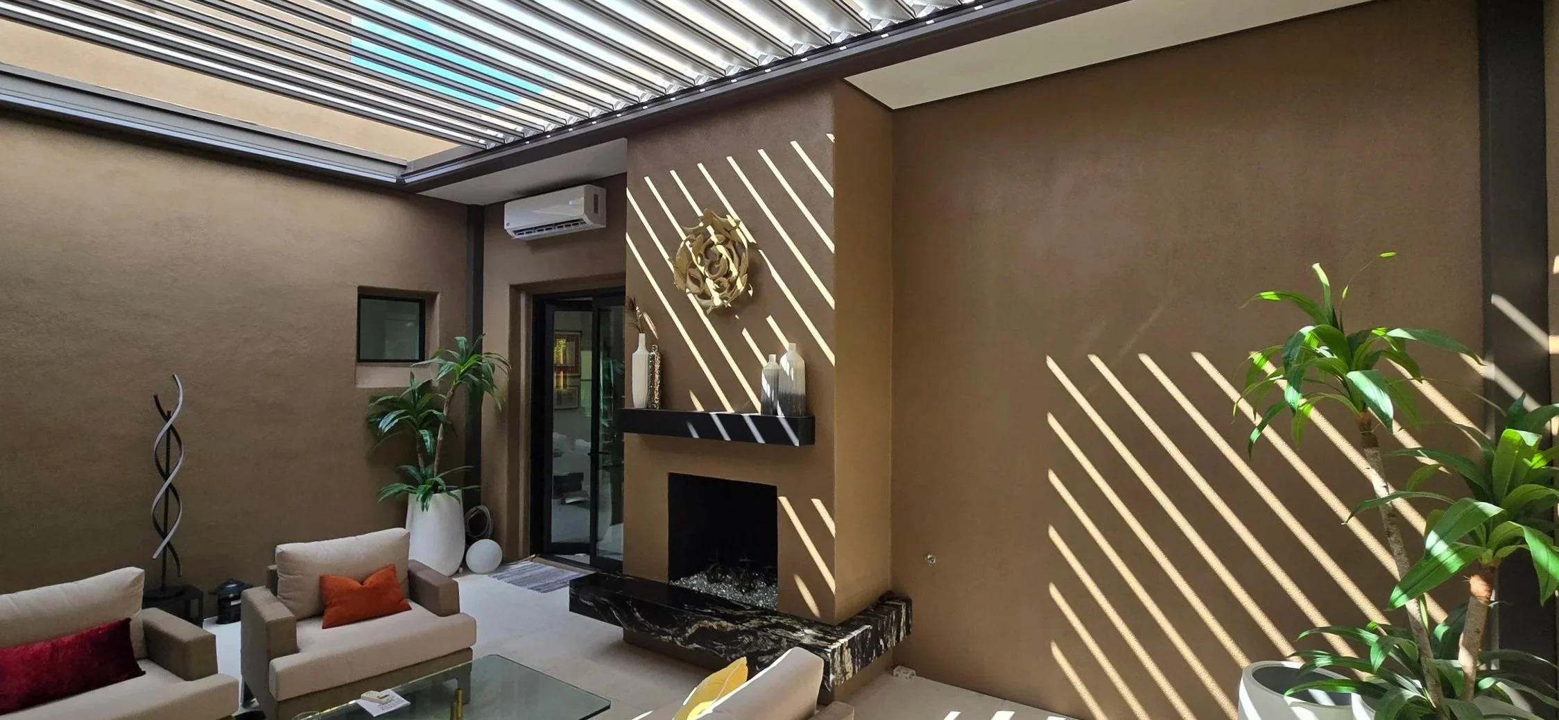 Modern fireplace feature beneath retractable louvered roof with shadow patterns on wall.