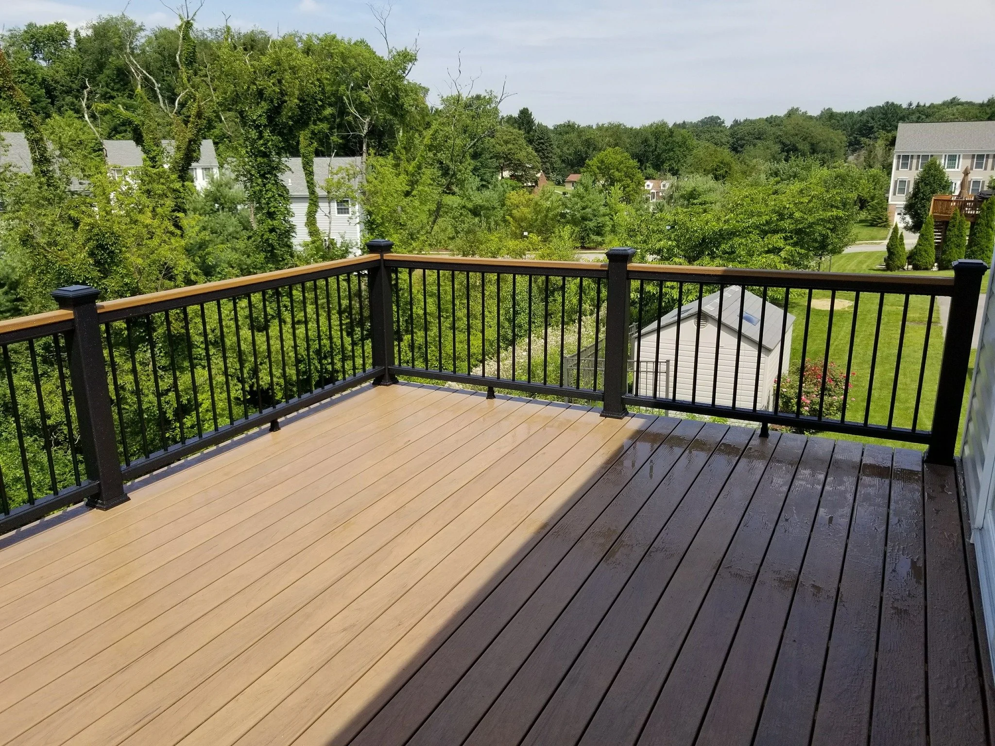 View from a wooden deck with black metal railing, overlooking green trees, neighboring houses, and a clear sky.