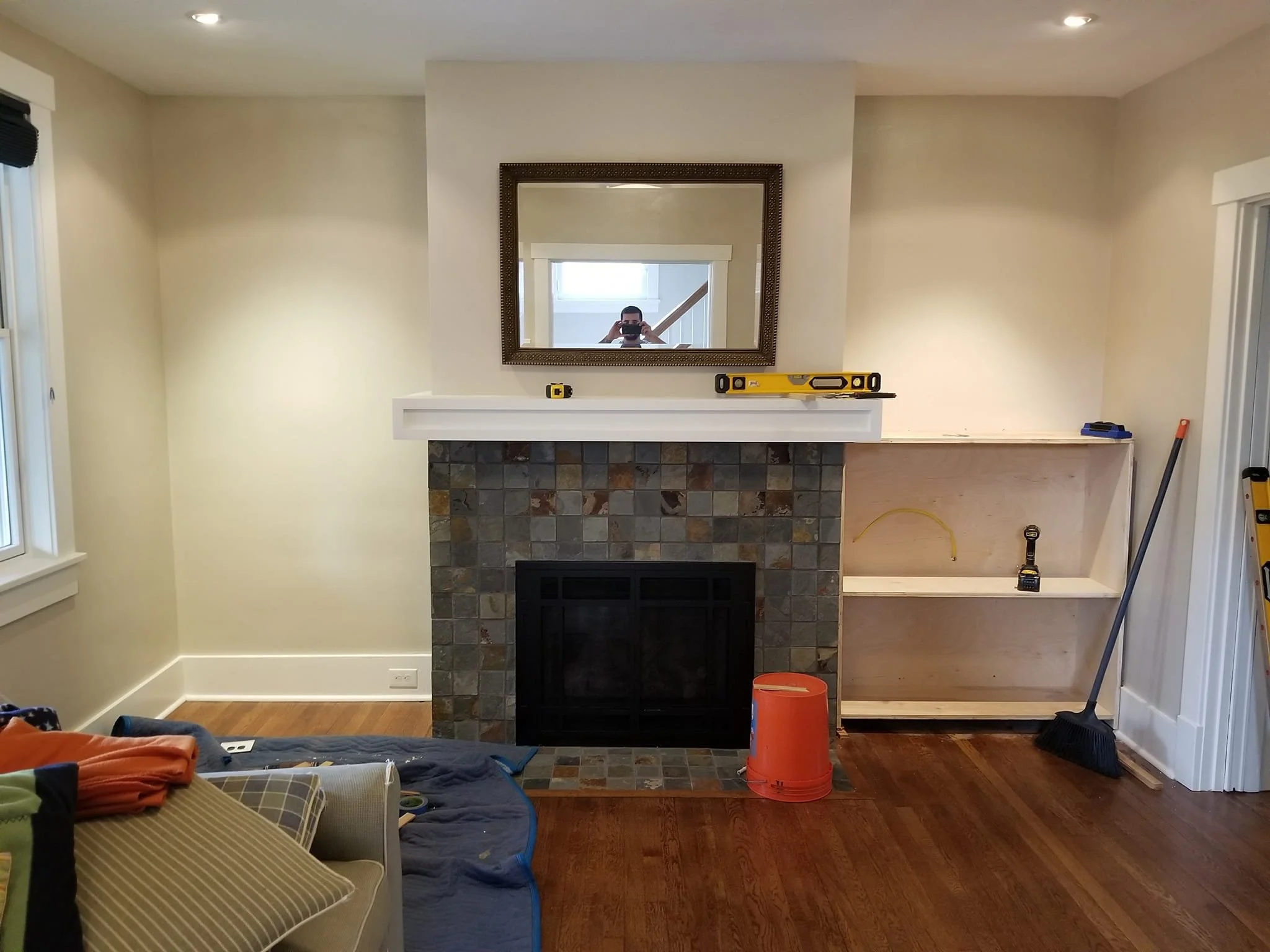 Living room fireplace under renovation with tools and supplies. The fireplace has a slate tile front and a white mantel, with a large mirror hanging above. Construction materials, including a level, a yellow tape measure, a cordless drill, and a broo