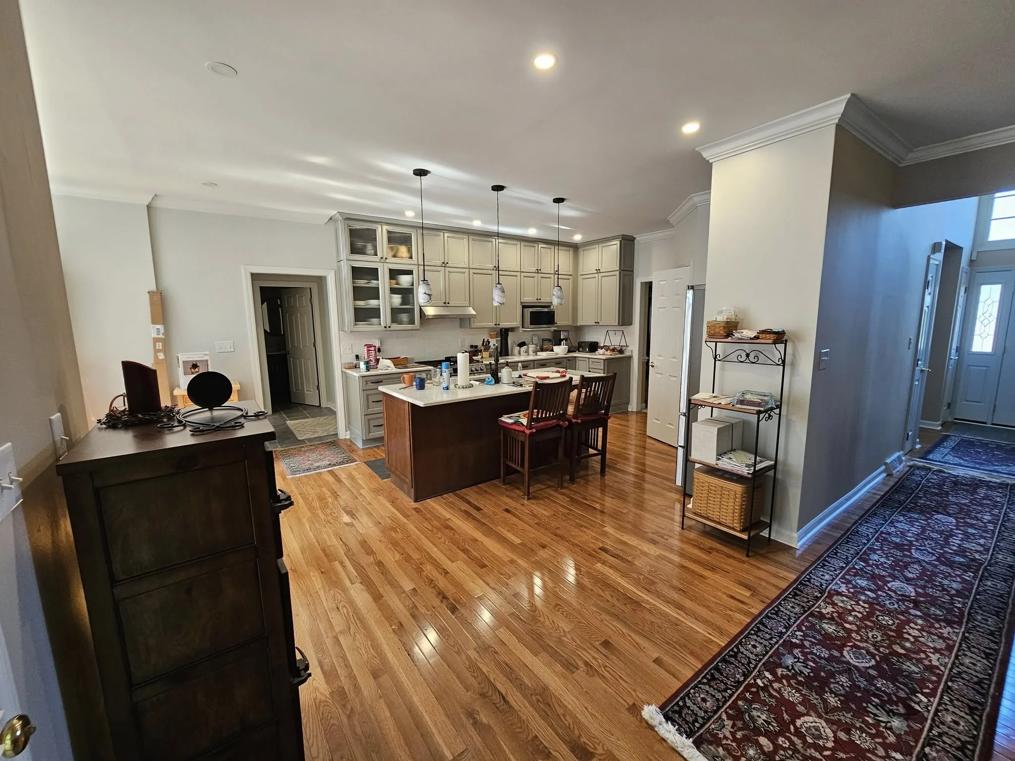 Spacious kitchen with gray cabinets, stainless steel appliances, a central island, and hardwood floors. To the right, an entryway with a decorative rug and a hallway.