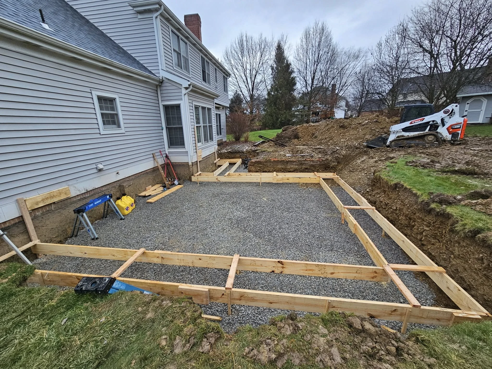 Construction site with wooden framing and gravel foundation for a new patio or extension, adjacent to a white house with blue siding, and a small excavator on a dirt mound in the background.