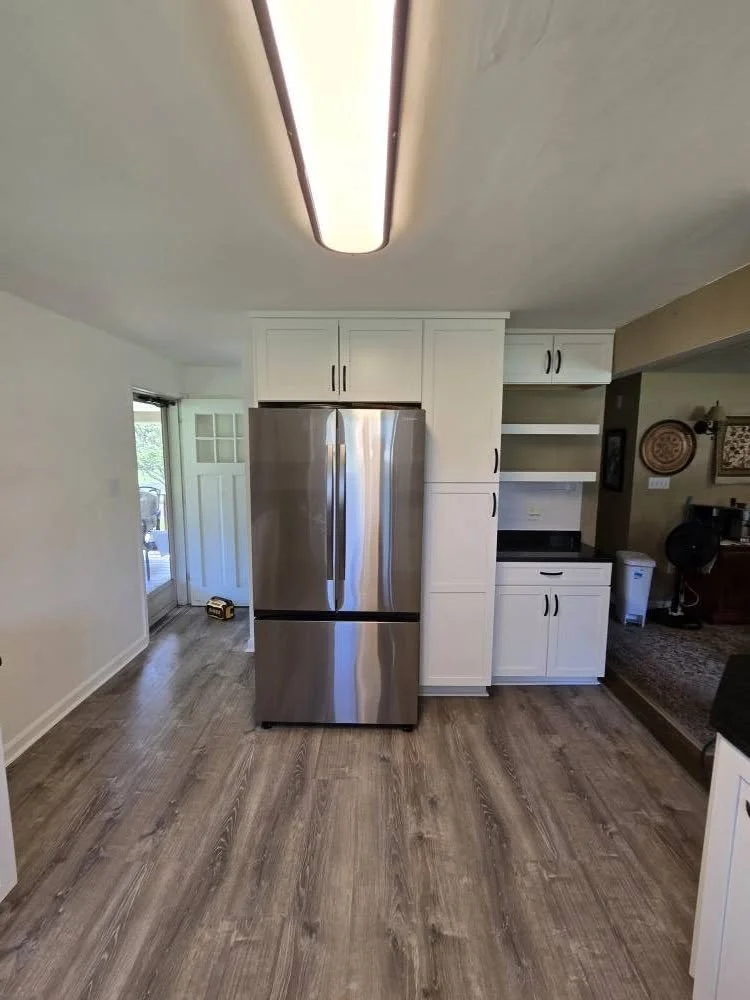 A modern kitchen with a stainless steel refrigerator, white cabinets, and wood flooring, viewed from the living area.
