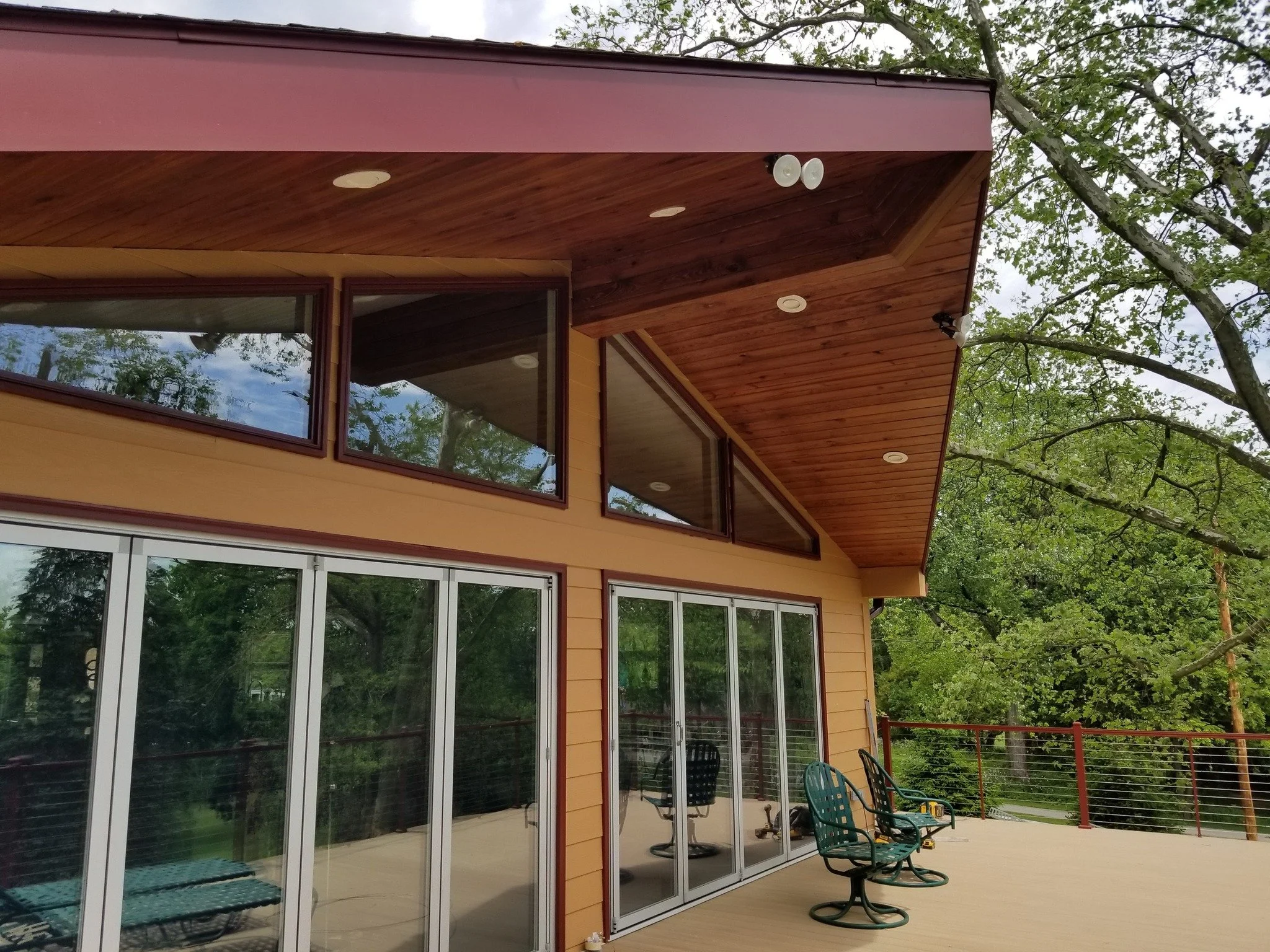 A house with large glass sliding doors and windows, a wooden overhang ceiling, and an outdoor deck with chairs, surrounded by green trees.
