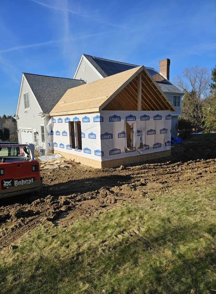A house under construction with new siding and roofing being installed, construction equipment in the yard, and a clear blue sky.
