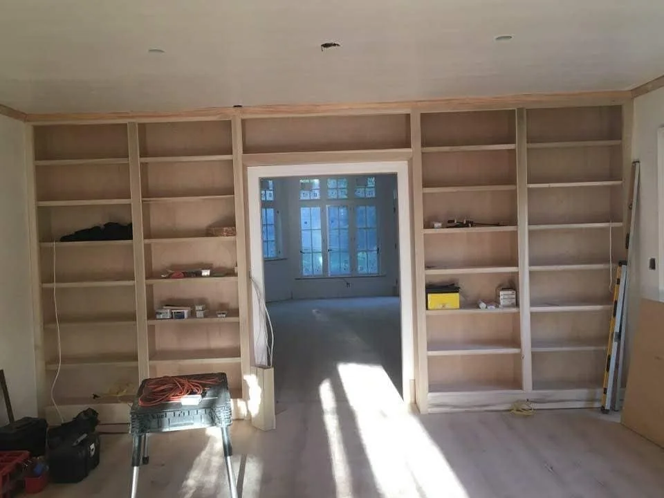 Room under construction with built-in wooden bookshelves, tools, and a ladder, leading to another room with large windows.